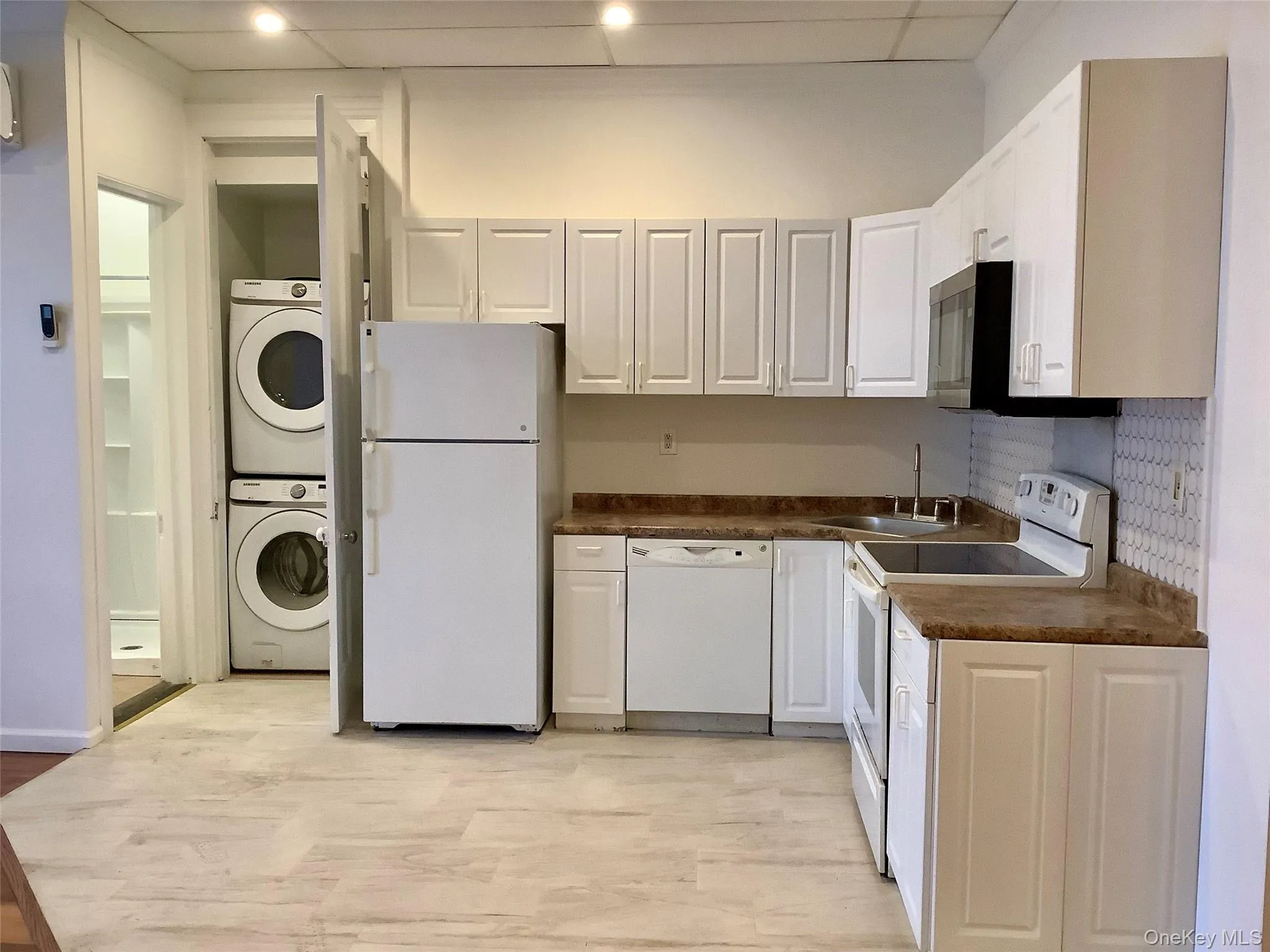 Kitchen with white appliances, white cabinetry, dark countertops, stacked washer / drying machine, and light wood-style floors Kitchen with white appliances, white cabinetry, dark countertops, stacked washer / drying machine, and light wood-style floors