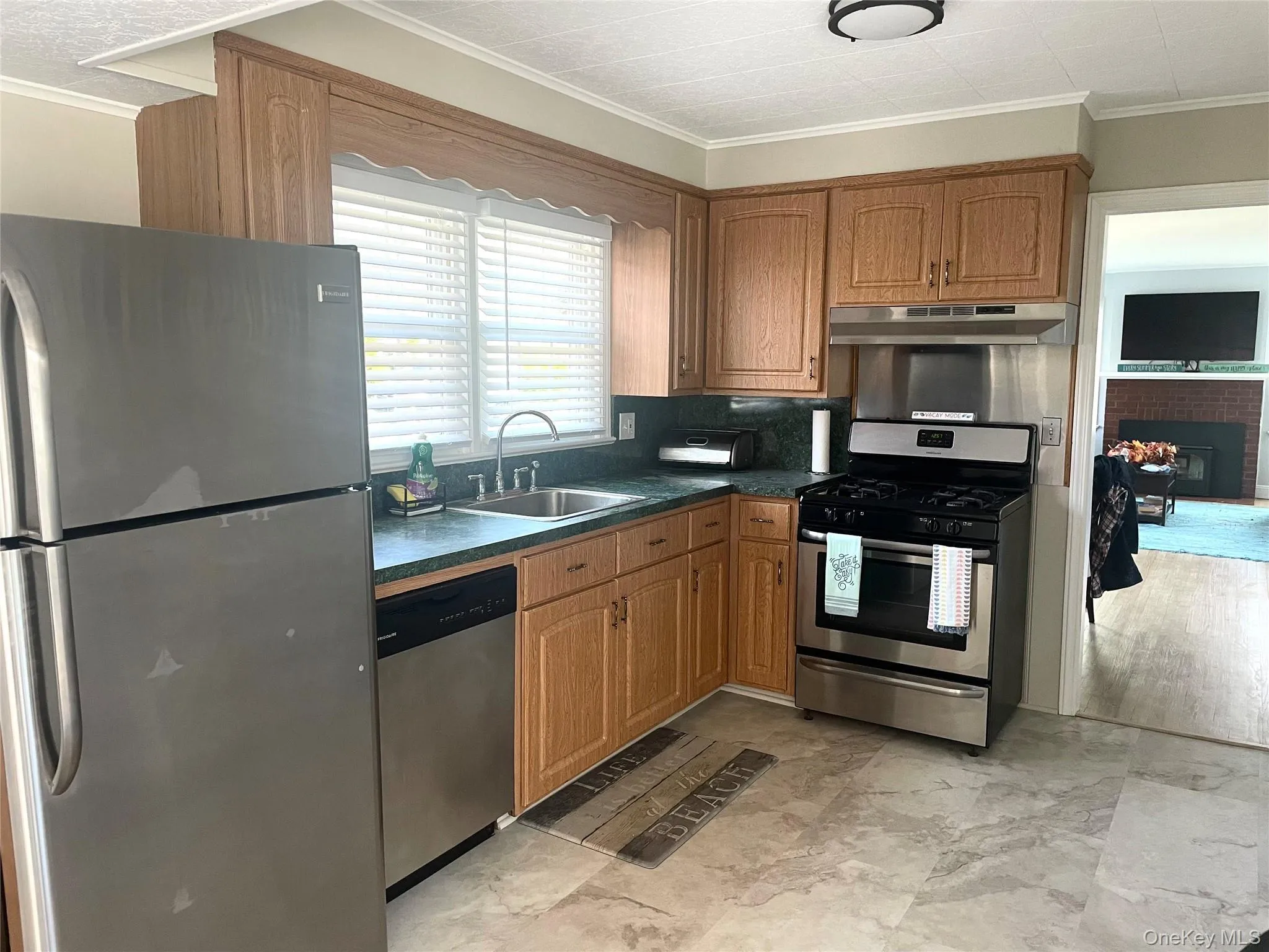 Kitchen with appliances with stainless steel finishes, brown cabinetry, dark countertops, ornamental molding, and under cabinet range hood Kitchen with appliances with stainless steel finishes, brown cabinetry, dark countertops, ornamental molding, and under cabinet range hood