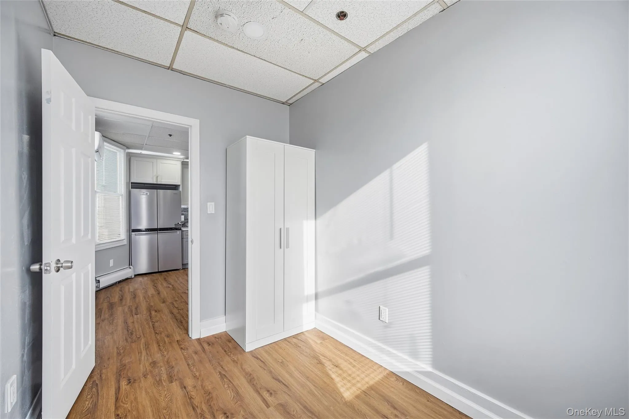 Unfurnished bedroom with a drop ceiling, freestanding refrigerator, a baseboard radiator, and wood finished floors Unfurnished bedroom with a drop ceiling, freestanding refrigerator, a baseboard radiator, and wood finished floors