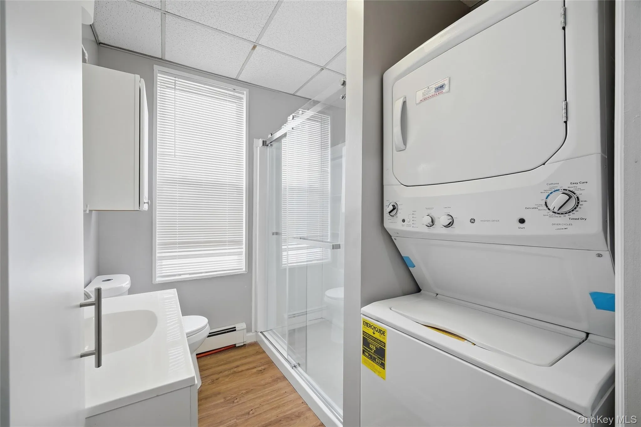 Laundry area with a drop ceiling, a baseboard radiator, stacked washing machine and dryer, healthy amount of natural light, and light wood finished floors Laundry area with a drop ceiling, a baseboard radiator, stacked washing machine and dryer, healthy amount of natural light, and light wood finished floors