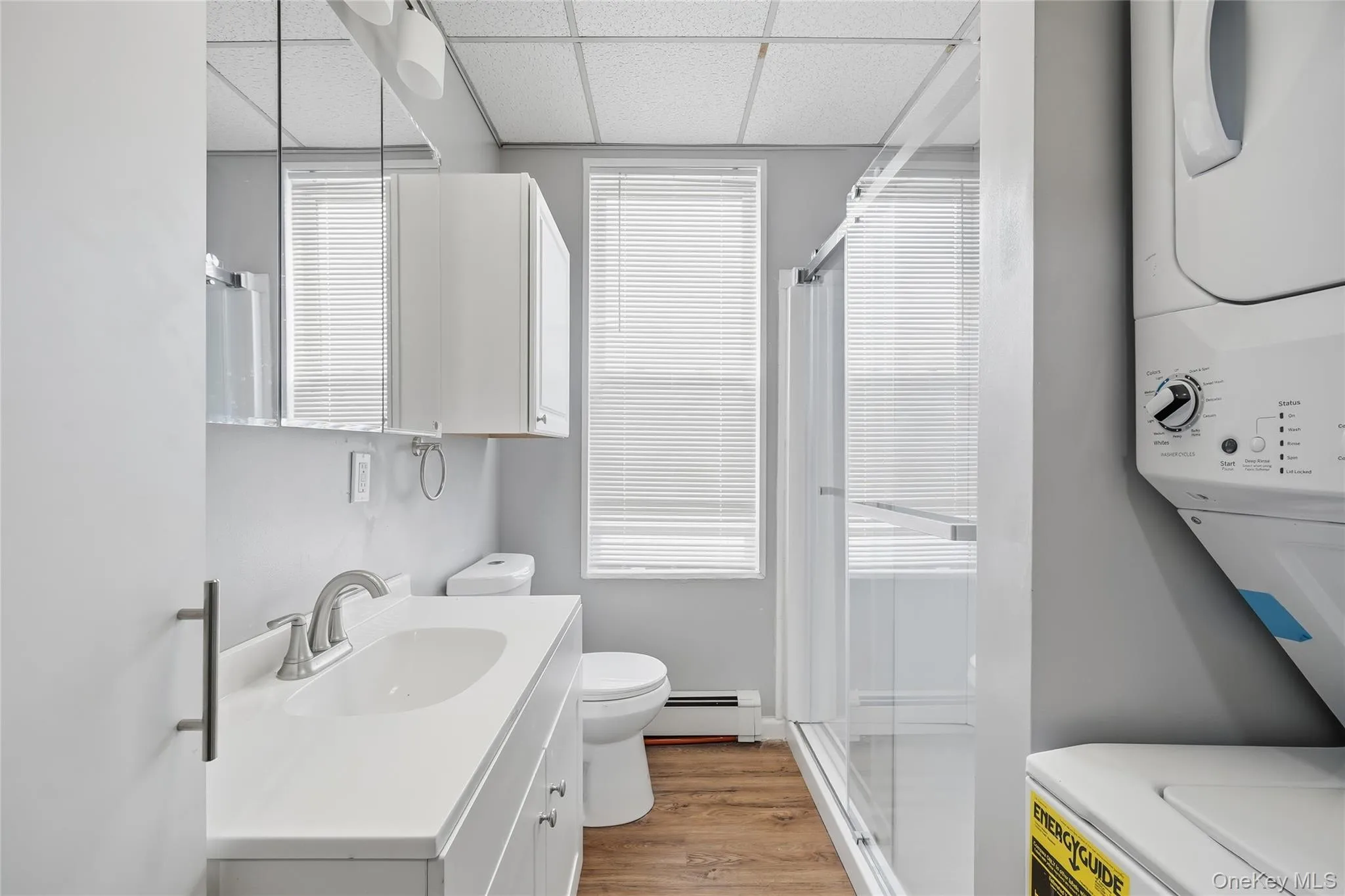 Full bath featuring a drop ceiling, a stall shower, vanity, stacked washer and clothes dryer, and a baseboard heating unit Full bath featuring a drop ceiling, a stall shower, vanity, stacked washer and clothes dryer, and a baseboard heating unit