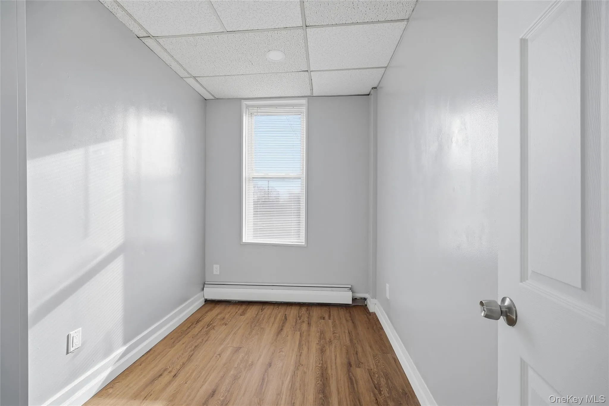 Empty room with a baseboard heating unit, a paneled ceiling, and wood finished floors Empty room with a baseboard heating unit, a paneled ceiling, and wood finished floors