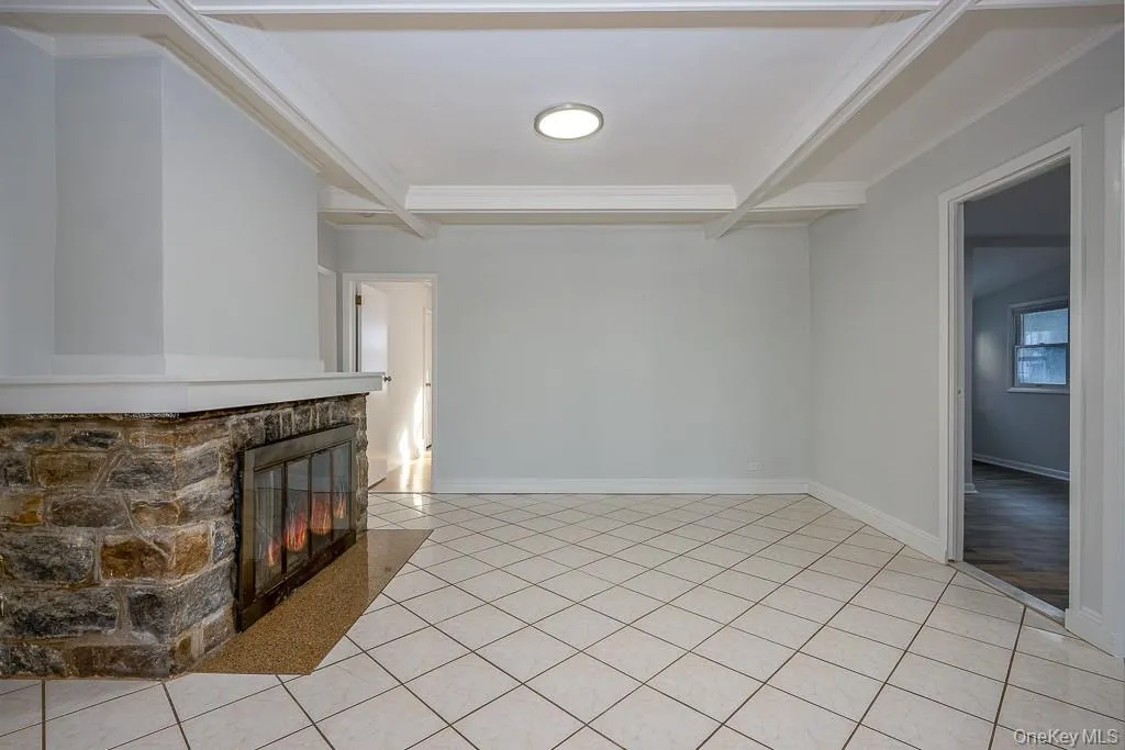 Unfurnished living room featuring a stone fireplace, beam ceiling, light tile patterned floors, and crown molding Unfurnished living room featuring a stone fireplace, beam ceiling, light tile patterned floors, and crown molding