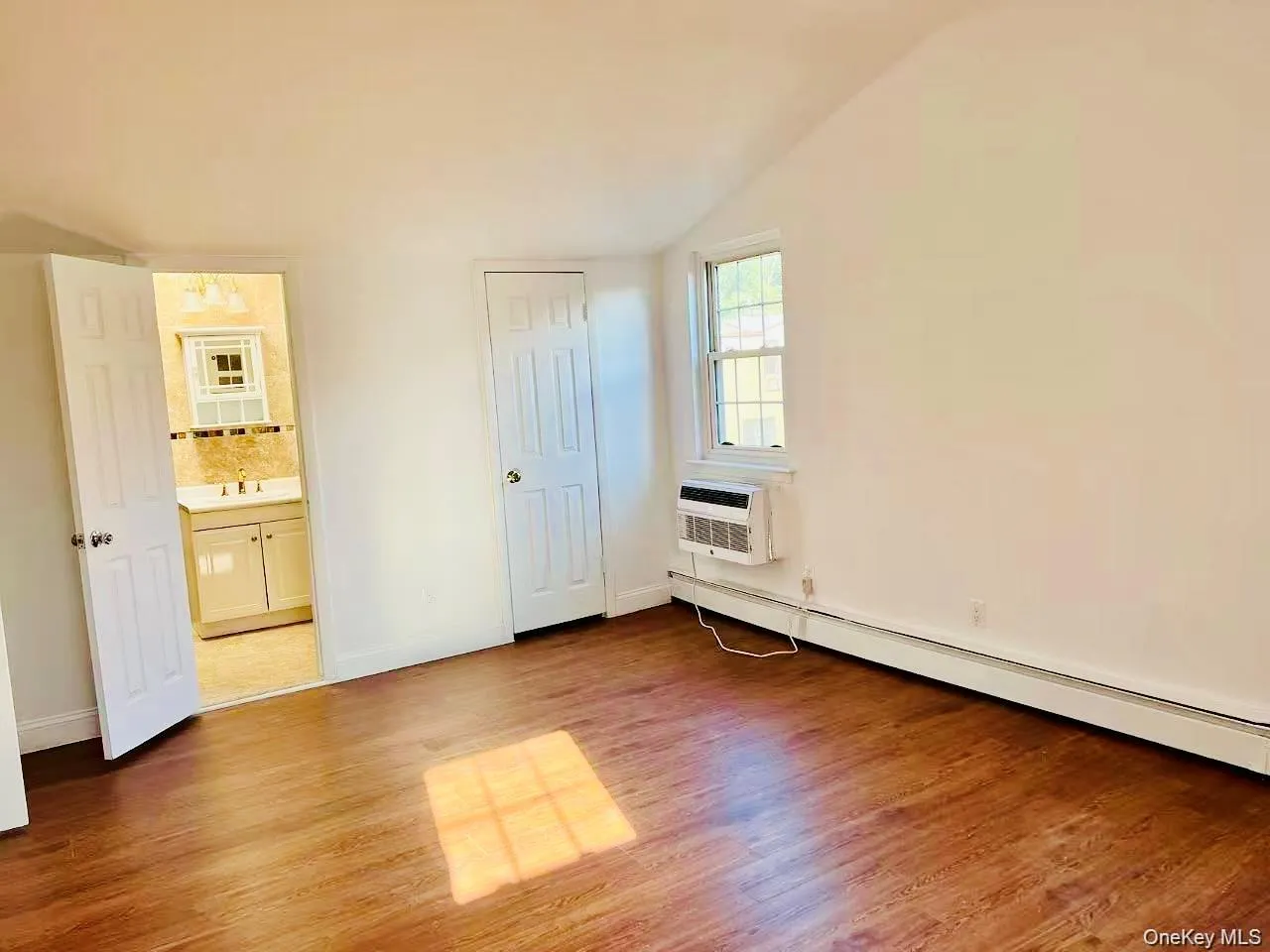 Unfurnished bedroom featuring baseboard heating, wood finished floors, a wall mounted air conditioner, vaulted ceiling, and connected bathroom Unfurnished bedroom featuring baseboard heating, wood finished floors, a wall mounted air conditioner, vaulted ceiling, and connected bathroom