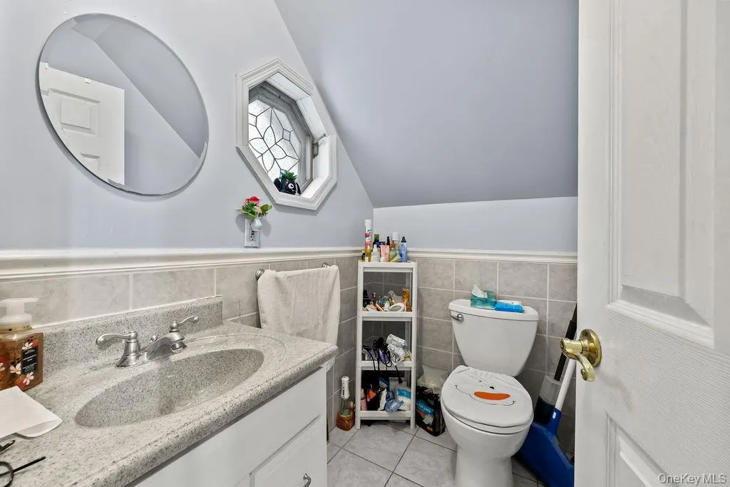 Half bathroom with vanity, a wainscoted wall, tile walls, light tile patterned flooring, and lofted ceiling Half bathroom with vanity, a wainscoted wall, tile walls, light tile patterned flooring, and lofted ceiling