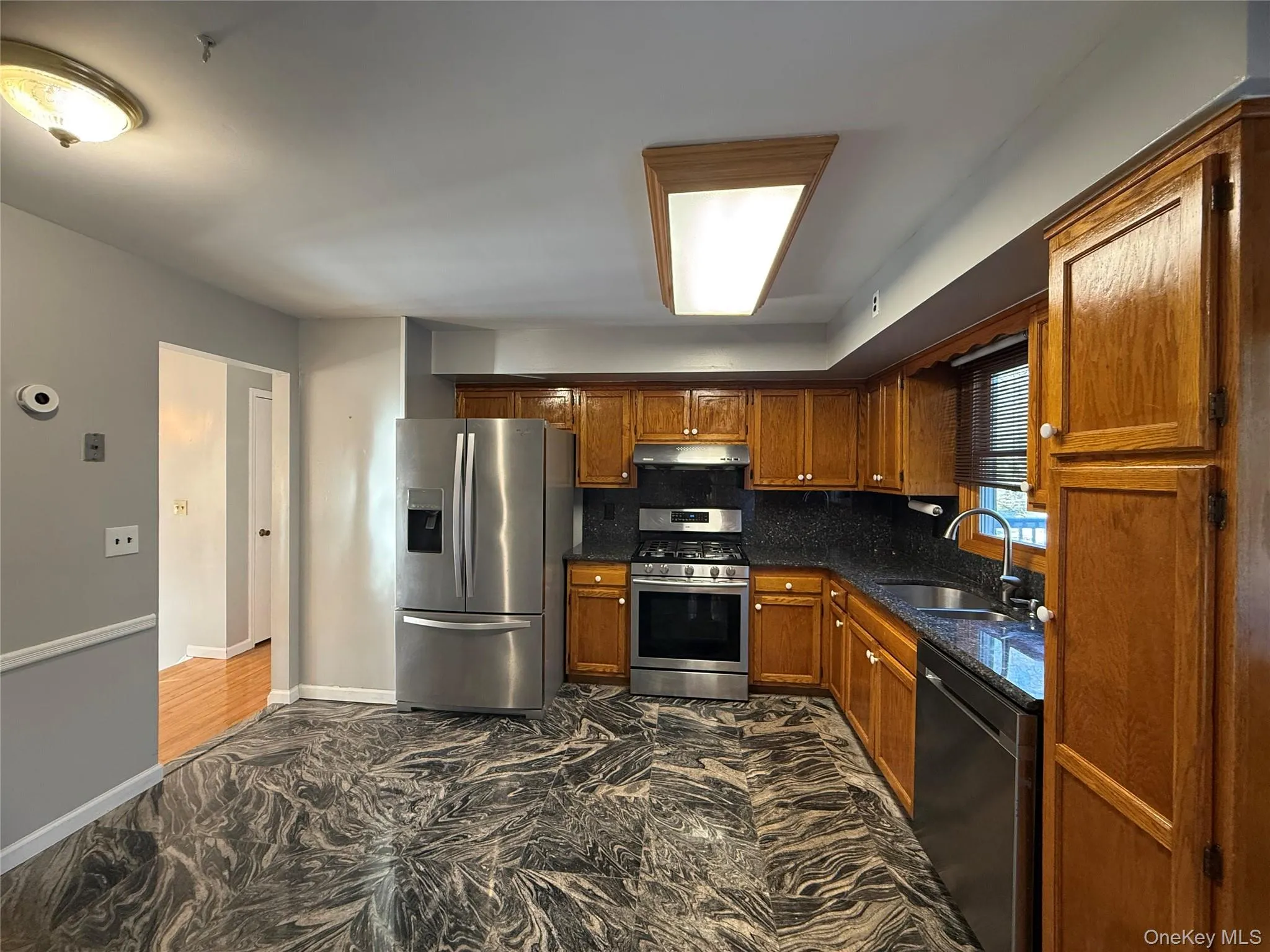 Kitchen with stainless steel appliances, dark marble finish floors, brown cabinetry, decorative backsplash, and dark stone counters Kitchen with stainless steel appliances, dark marble finish floors, brown cabinetry, decorative backsplash, and dark stone counters