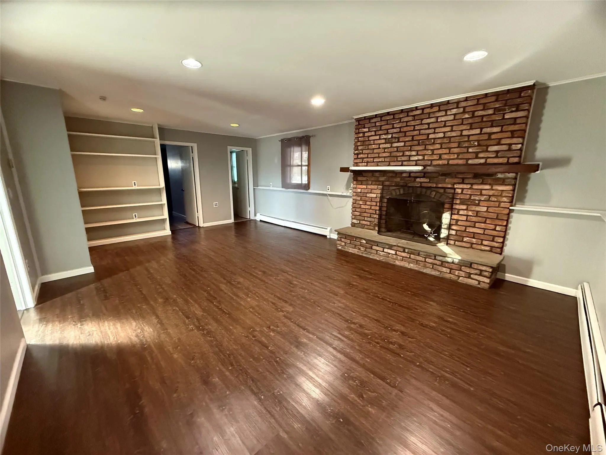 Unfurnished living room with baseboard heating, a brick fireplace, recessed lighting, dark wood-style floors, and ornamental molding Unfurnished living room with baseboard heating, a brick fireplace, recessed lighting, dark wood-style floors, and ornamental molding