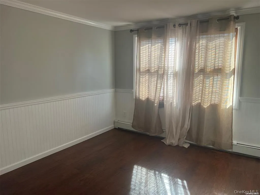 Empty room featuring wainscoting, dark wood-style flooring, ornamental molding, and a baseboard radiator Empty room featuring wainscoting, dark wood-style flooring, ornamental molding, and a baseboard radiator