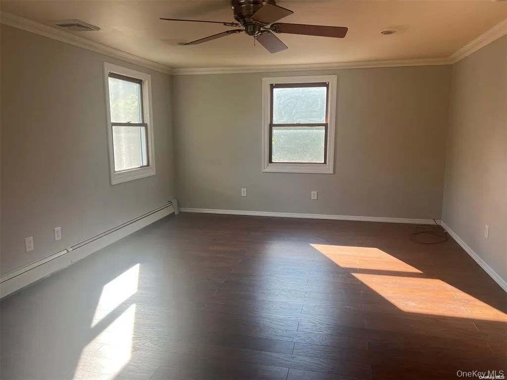 Spare room featuring crown molding, dark wood finished floors, healthy amount of natural light, a ceiling fan, and a baseboard heating unit Spare room featuring crown molding, dark wood finished floors, healthy amount of natural light, a ceiling fan, and a baseboard heating unit