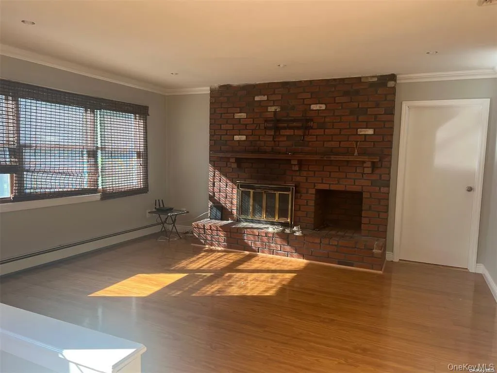 Unfurnished living room with a brick fireplace, wood finished floors, ornamental molding, and baseboard heating Unfurnished living room with a brick fireplace, wood finished floors, ornamental molding, and baseboard heating