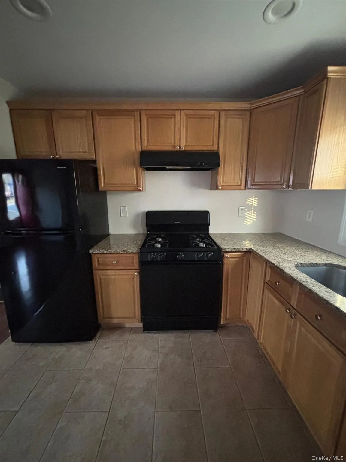 Kitchen featuring black appliances, light stone countertops, under cabinet range hood, dark tile patterned floors, and brown cabinets Kitchen featuring black appliances, light stone countertops, under cabinet range hood, dark tile patterned floors, and brown cabinets