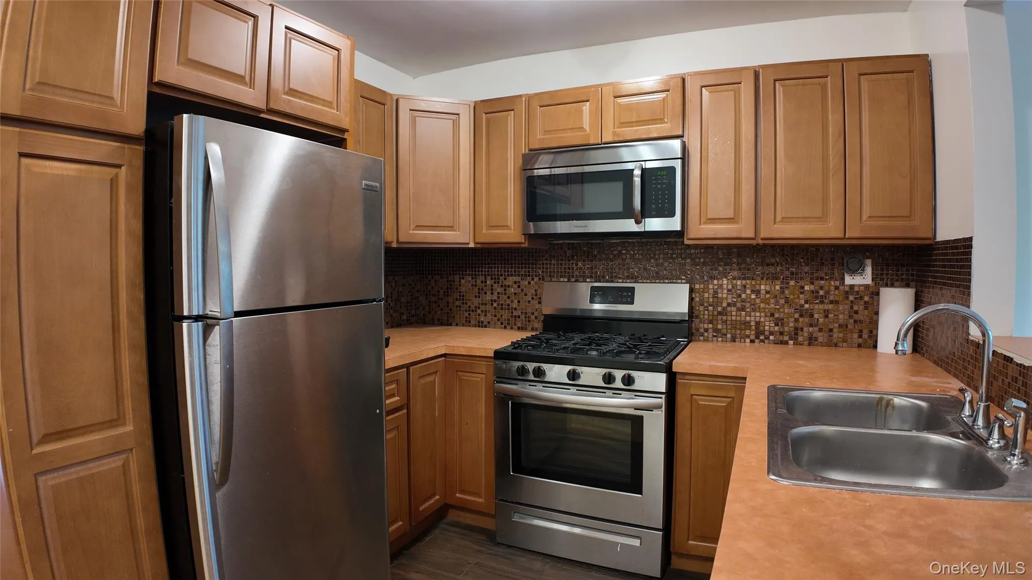 Kitchen featuring appliances with stainless steel finishes, light countertops, dark wood-type flooring, tasteful backsplash, and brown cabinets Kitchen featuring appliances with stainless steel finishes, light countertops, dark wood-type flooring, tasteful backsplash, and brown cabinets