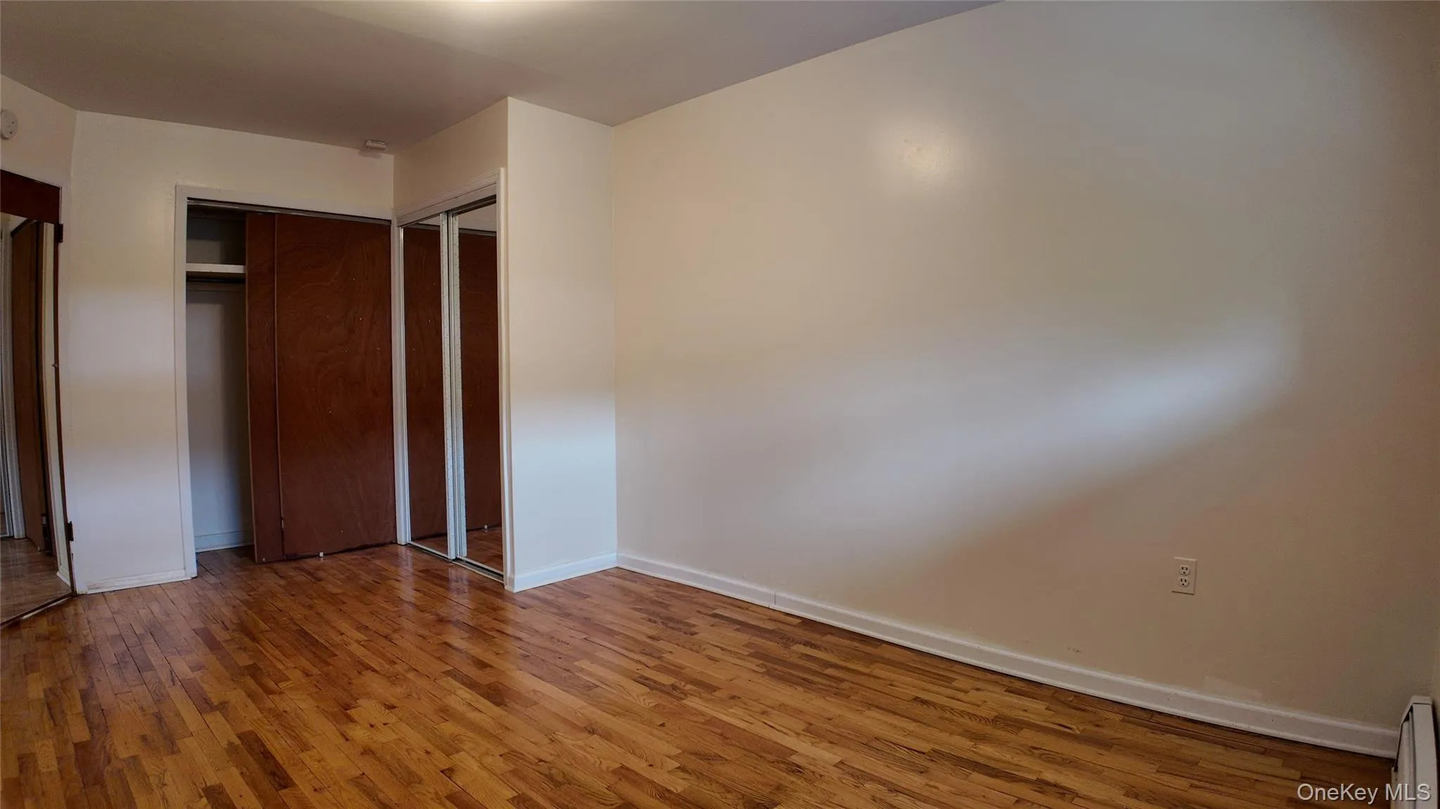 Unfurnished bedroom with wood finished floors, a baseboard heating unit, and two closets Unfurnished bedroom with wood finished floors, a baseboard heating unit, and two closets