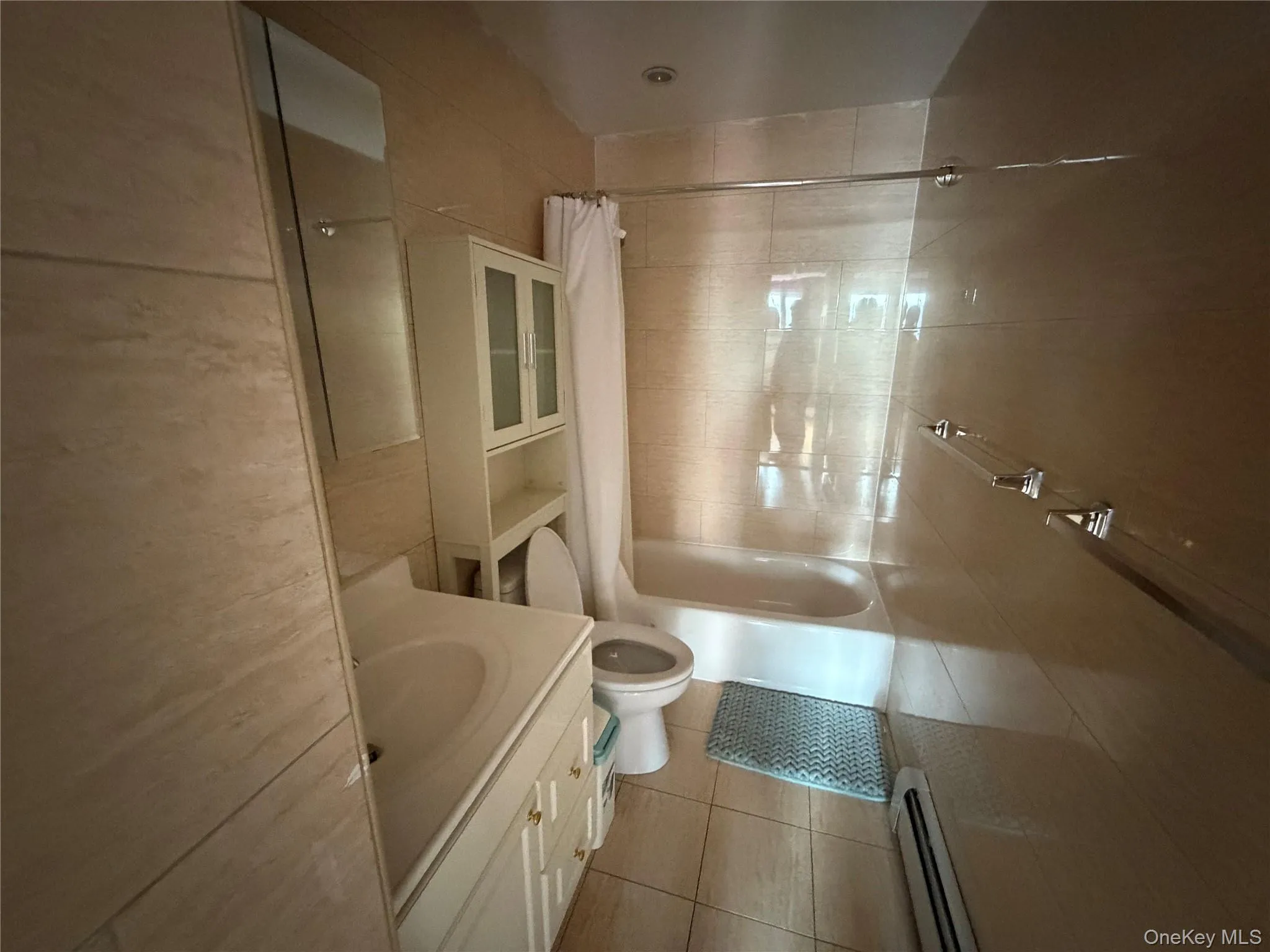 Full bath with vanity, baseboard heating, shower / bath combo with shower curtain, light tile patterned floors, and tile walls Full bath with vanity, baseboard heating, shower / bath combo with shower curtain, light tile patterned floors, and tile walls