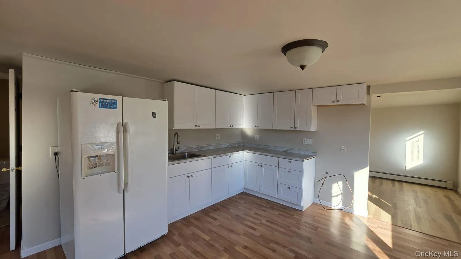 Kitchen with white fridge with ice dispenser, white cabinetry, dark wood-style flooring, a baseboard radiator, and light countertops Kitchen with white fridge with ice dispenser, white cabinetry, dark wood-style flooring, a baseboard radiator, and light countertops