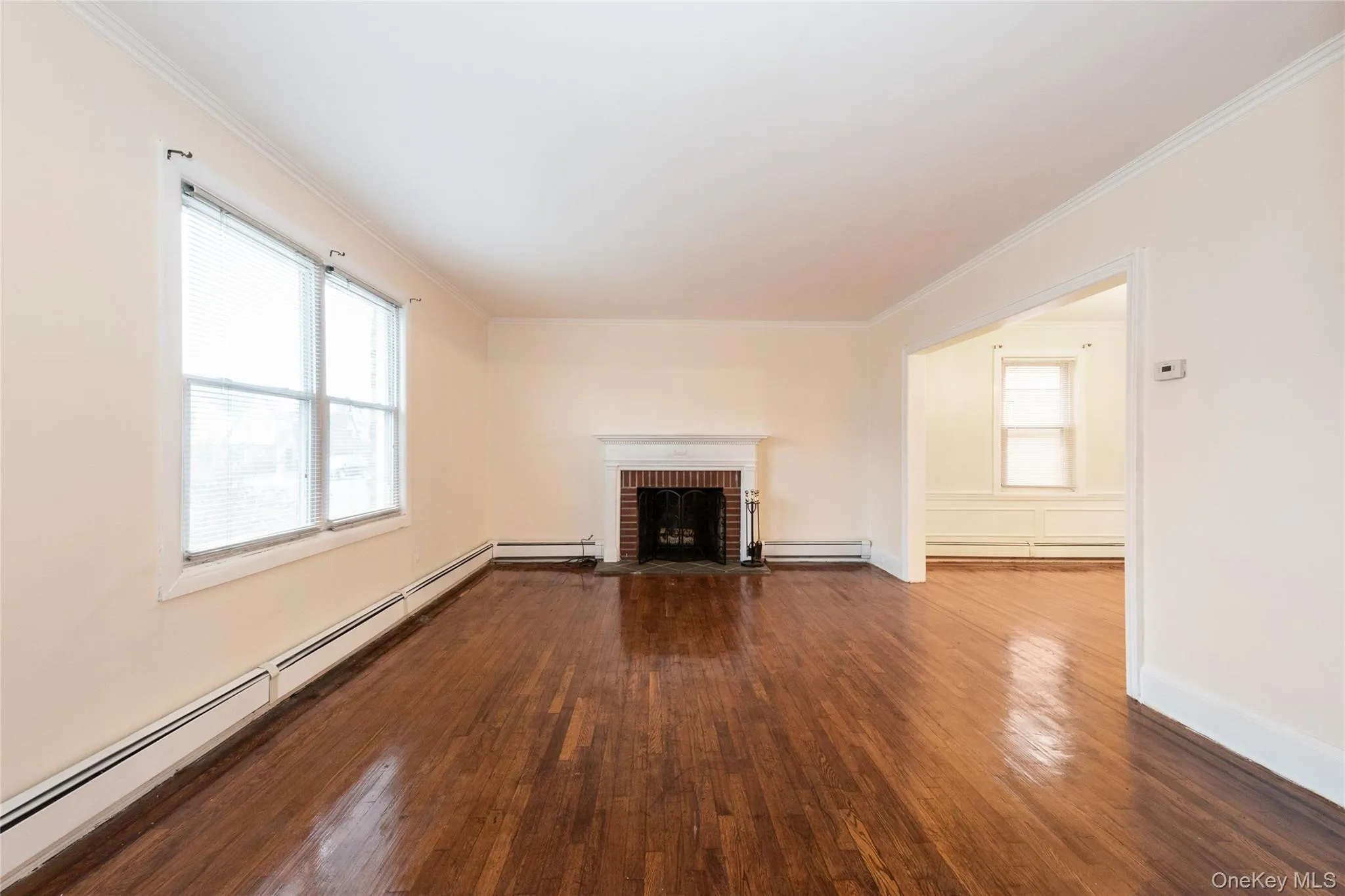 Unfurnished living room with baseboard heating, hardwood finished floors, crown molding, healthy amount of natural light, and a brick fireplace. Unfurnished living room with baseboard heating, hardwood finished floors, crown molding, healthy amount of natural light, and a brick fireplace.