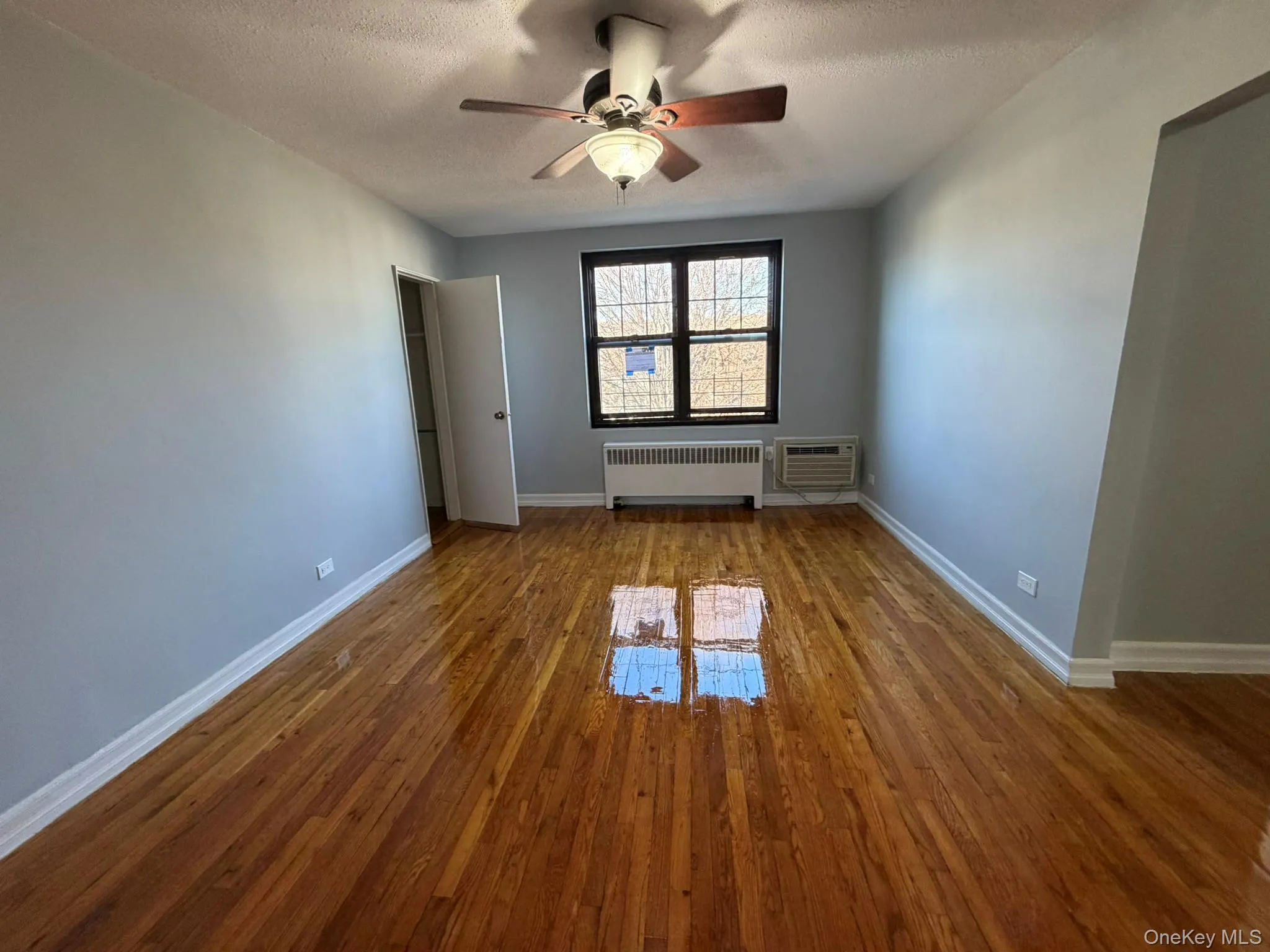 Unfurnished room with a textured ceiling, hardwood / wood-style flooring, radiator heating unit, a ceiling fan, and an AC wall unit Unfurnished room with a textured ceiling, hardwood / wood-style flooring, radiator heating unit, a ceiling fan, and an AC wall unit