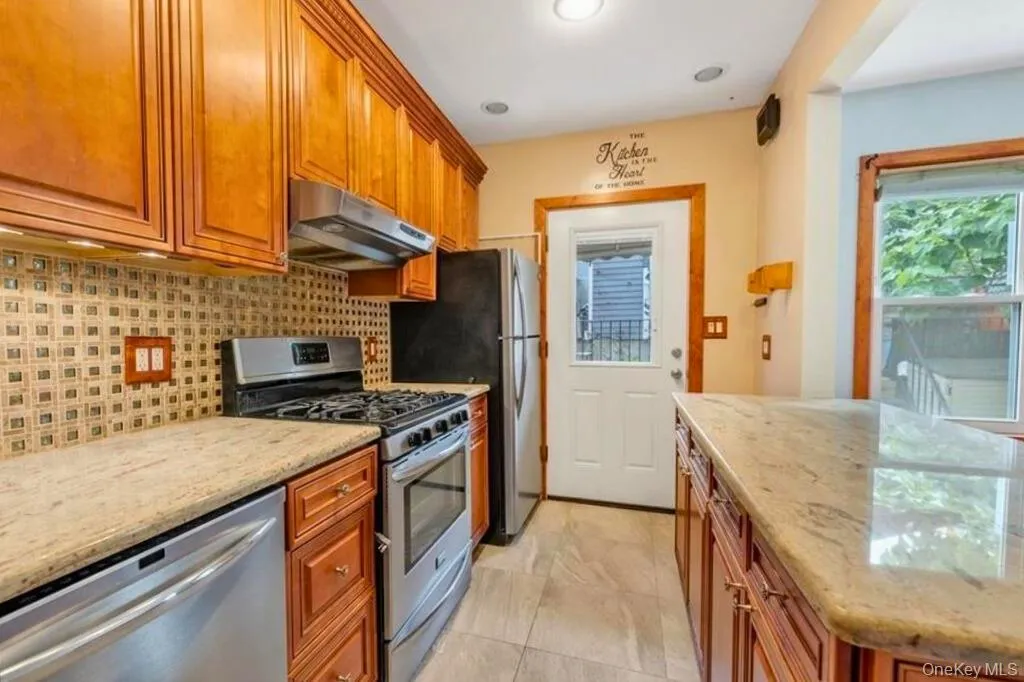 Kitchen with stainless steel appliances, light stone counters, a center island, brown cabinetry, and under cabinet range hood Kitchen with stainless steel appliances, light stone counters, a center island, brown cabinetry, and under cabinet range hood
