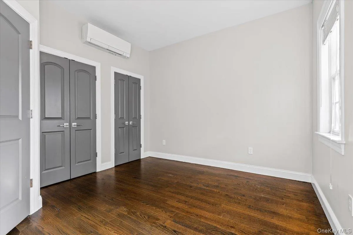Unfurnished bedroom with an AC wall unit, two closets, dark wood finished floors, and multiple windows Unfurnished bedroom with an AC wall unit, two closets, dark wood finished floors, and multiple windows