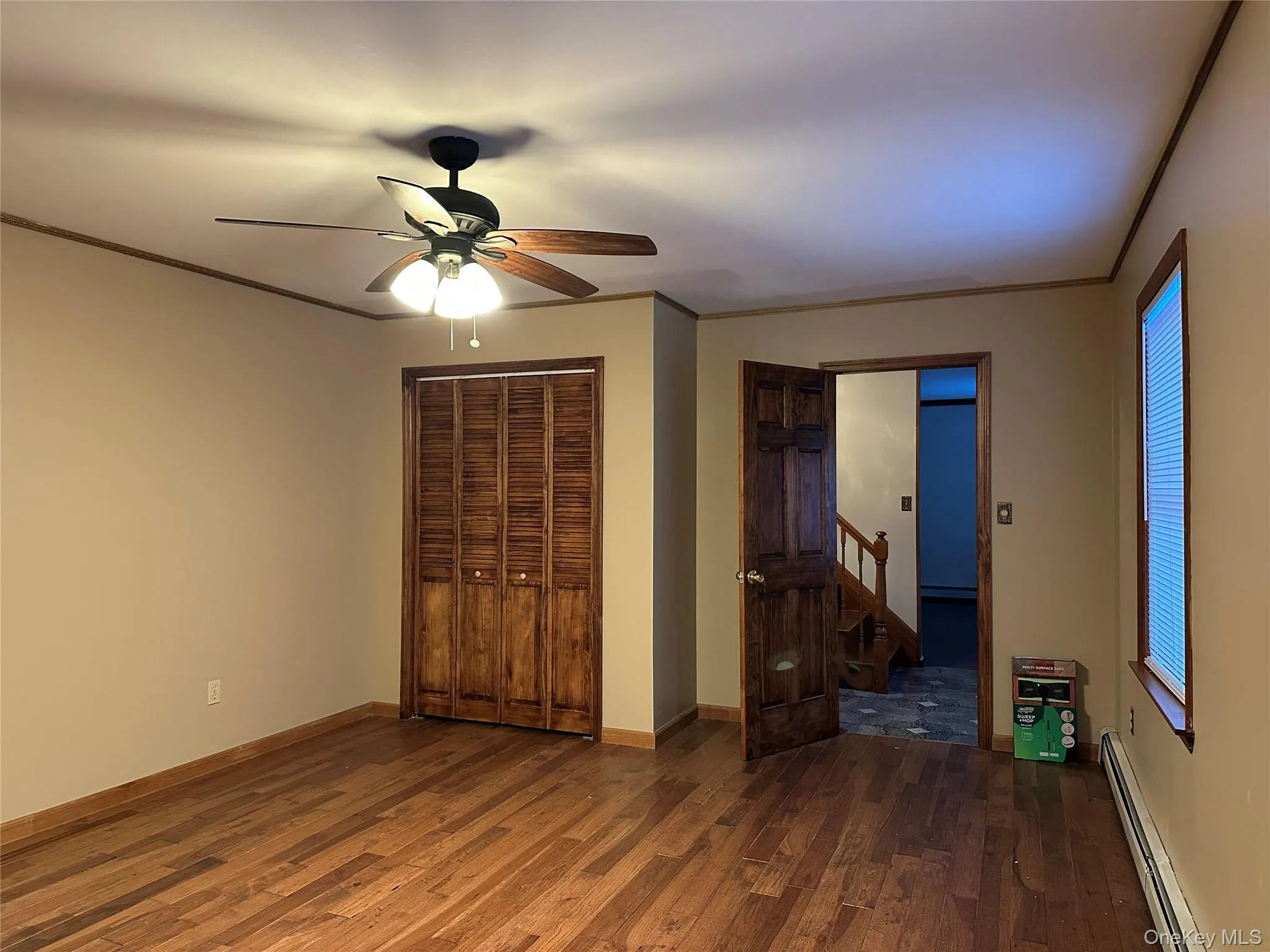 Unfurnished bedroom with baseboard heating, dark wood-style flooring, ceiling fan, a closet, and ornamental molding Unfurnished bedroom with baseboard heating, dark wood-style flooring, ceiling fan, a closet, and ornamental molding