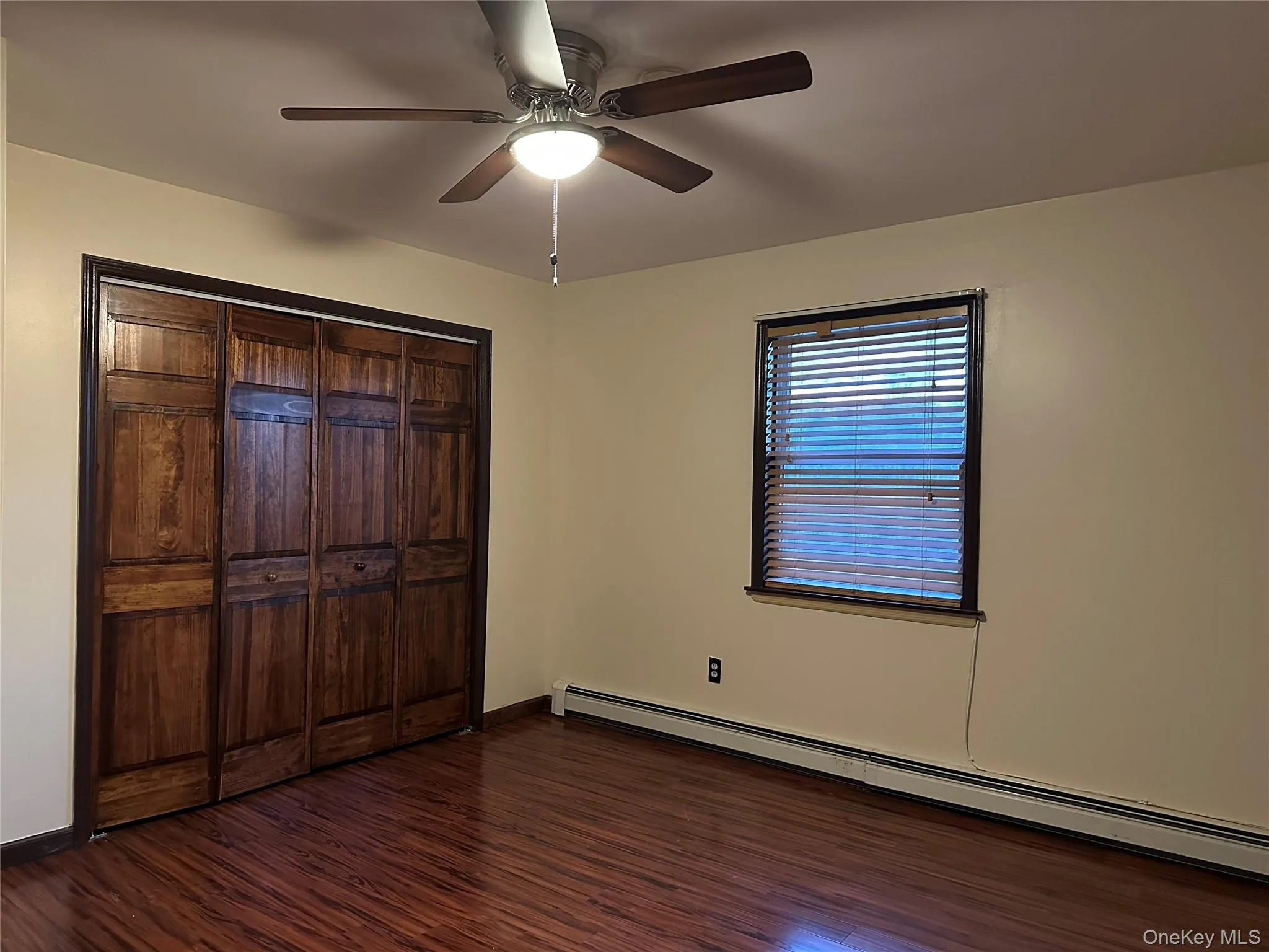 Unfurnished bedroom with a baseboard radiator, a ceiling fan, dark wood-type flooring, and a closet Unfurnished bedroom with a baseboard radiator, a ceiling fan, dark wood-type flooring, and a closet