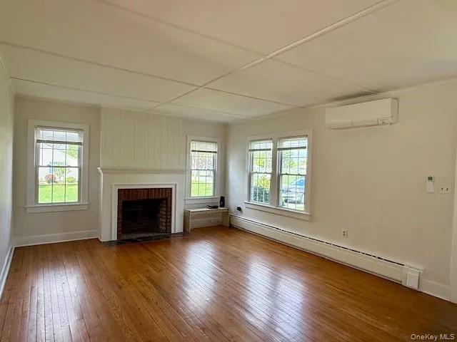 Unfurnished living room featuring a baseboard heating unit, a brick fireplace, hardwood / wood-style flooring, and plenty of natural light Unfurnished living room featuring a baseboard heating unit, a brick fireplace, hardwood / wood-style flooring, and plenty of natural light