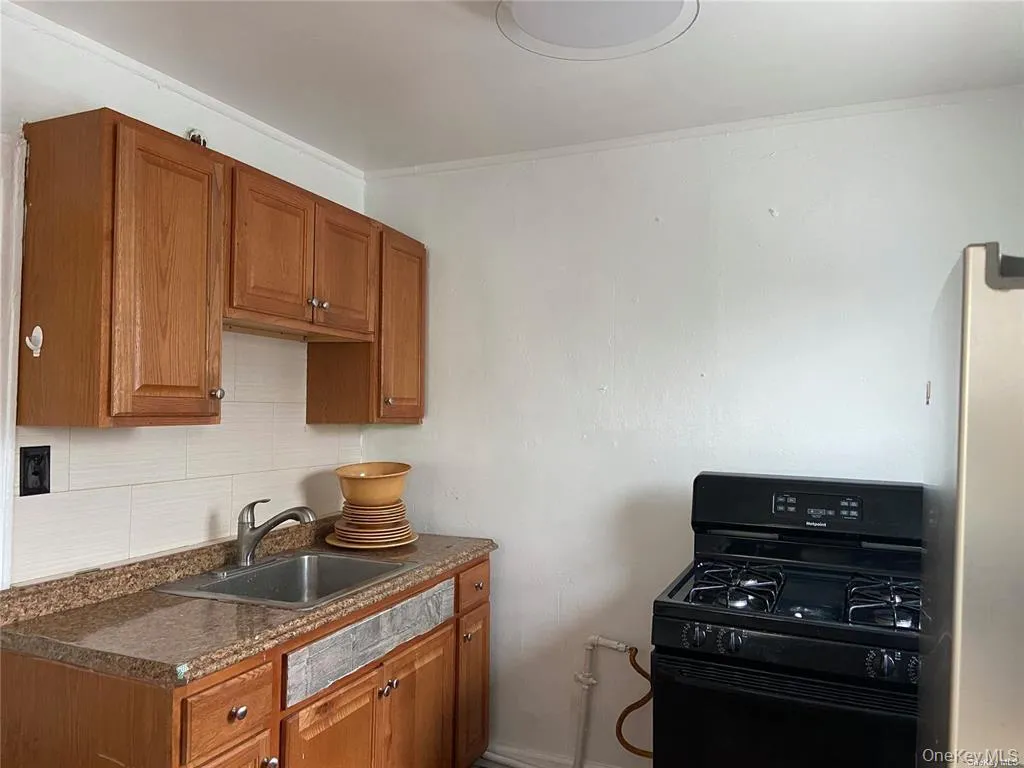 Kitchen with black gas range oven, brown cabinets, dark countertops, freestanding refrigerator, and tasteful backsplash Kitchen with black gas range oven, brown cabinets, dark countertops, freestanding refrigerator, and tasteful backsplash