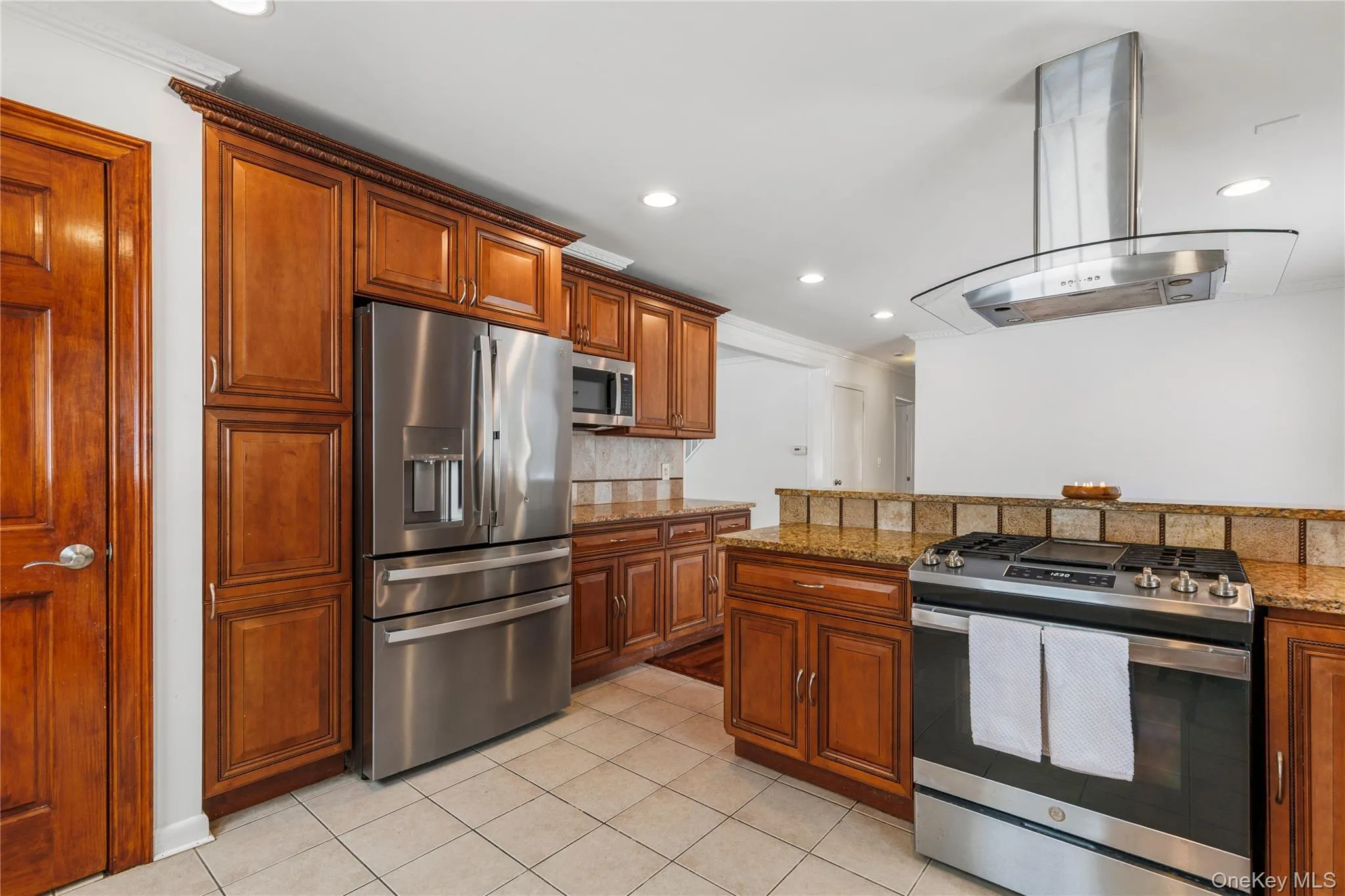 Kitchen featuring stainless steel appliances, light stone countertops, island range hood, brown cabinetry, and tasteful backsplash Kitchen featuring stainless steel appliances, light stone countertops, island range hood, brown cabinetry, and tasteful backsplash