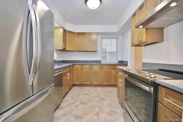Kitchen with appliances with stainless steel finishes, crown molding, under cabinet range hood, brown cabinetry, and dark countertops Kitchen with appliances with stainless steel finishes, crown molding, under cabinet range hood, brown cabinetry, and dark countertops