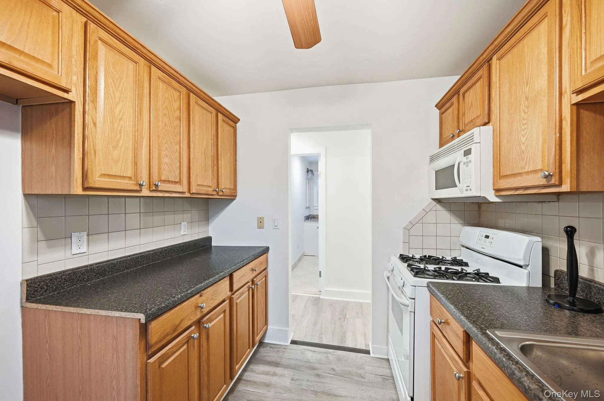 Kitchen featuring white appliances, dark countertops, decorative backsplash, brown cabinetry, and a ceiling fan Kitchen featuring white appliances, dark countertops, decorative backsplash, brown cabinetry, and a ceiling fan