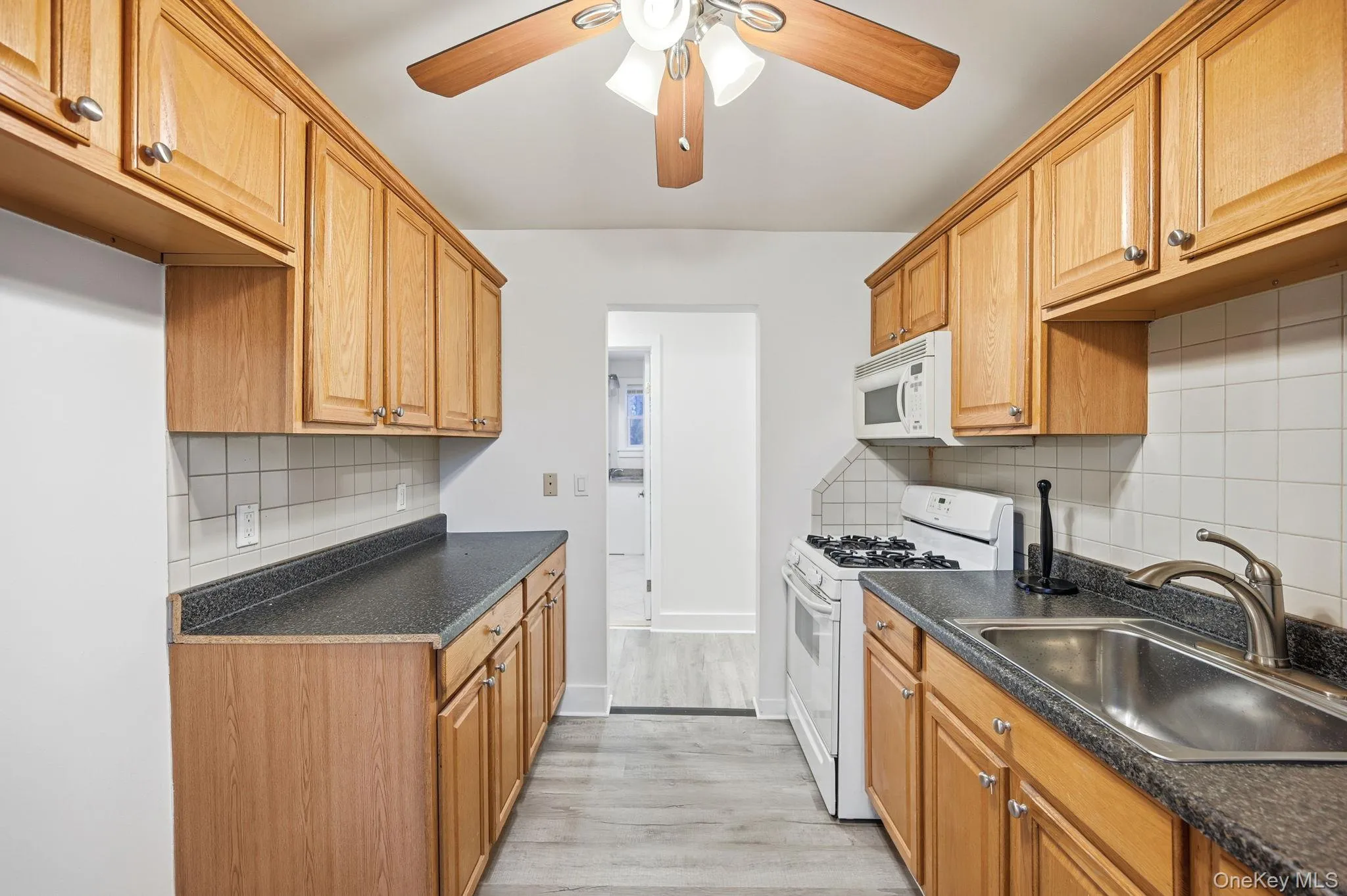 Kitchen featuring white appliances, a ceiling fan, dark countertops, light wood-style flooring, and tasteful backsplash Kitchen featuring white appliances, a ceiling fan, dark countertops, light wood-style flooring, and tasteful backsplash
