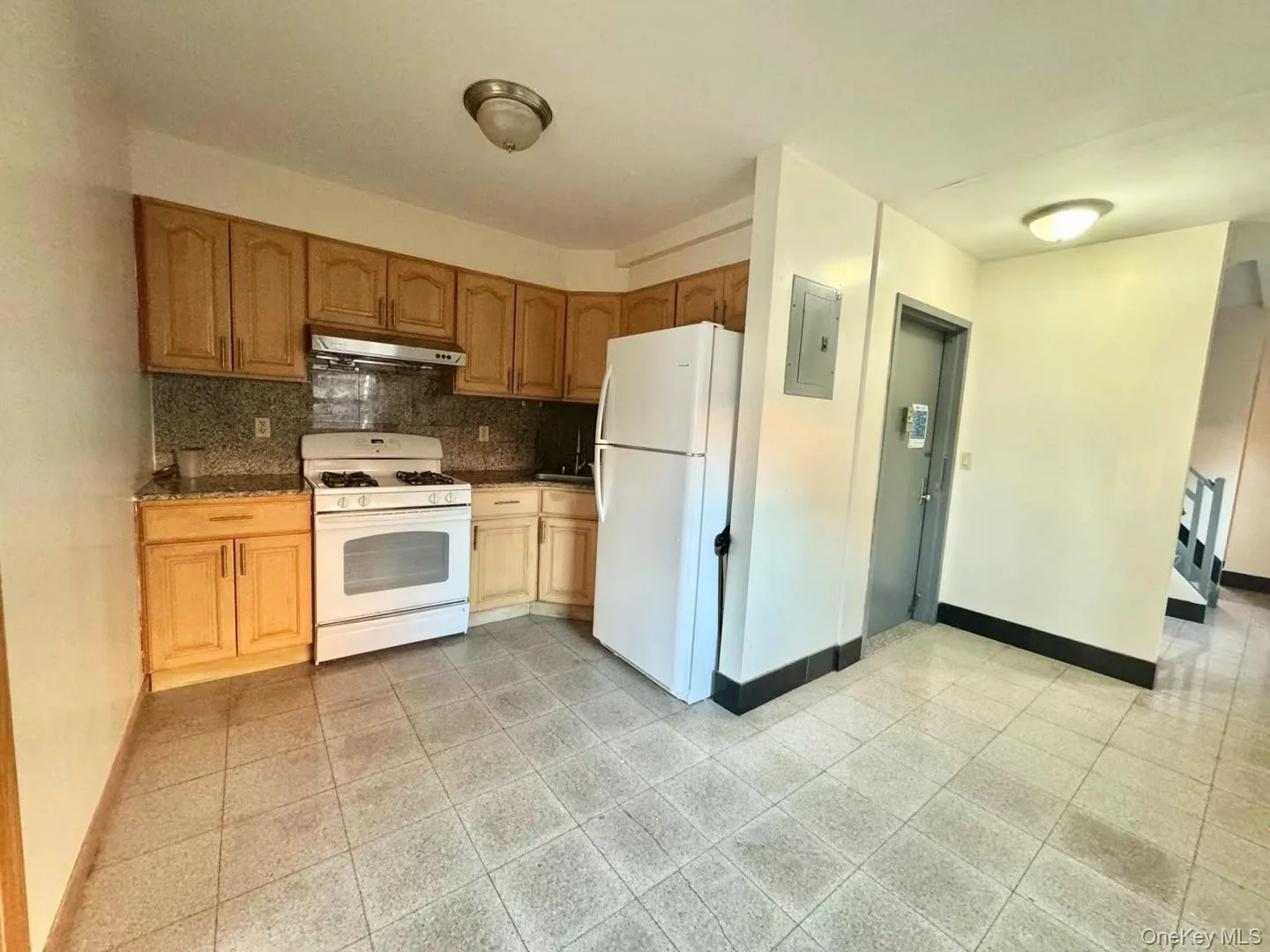 Kitchen featuring white appliances, under cabinet range hood, decorative backsplash, electric panel, and dark stone counters Kitchen featuring white appliances, under cabinet range hood, decorative backsplash, electric panel, and dark stone counters