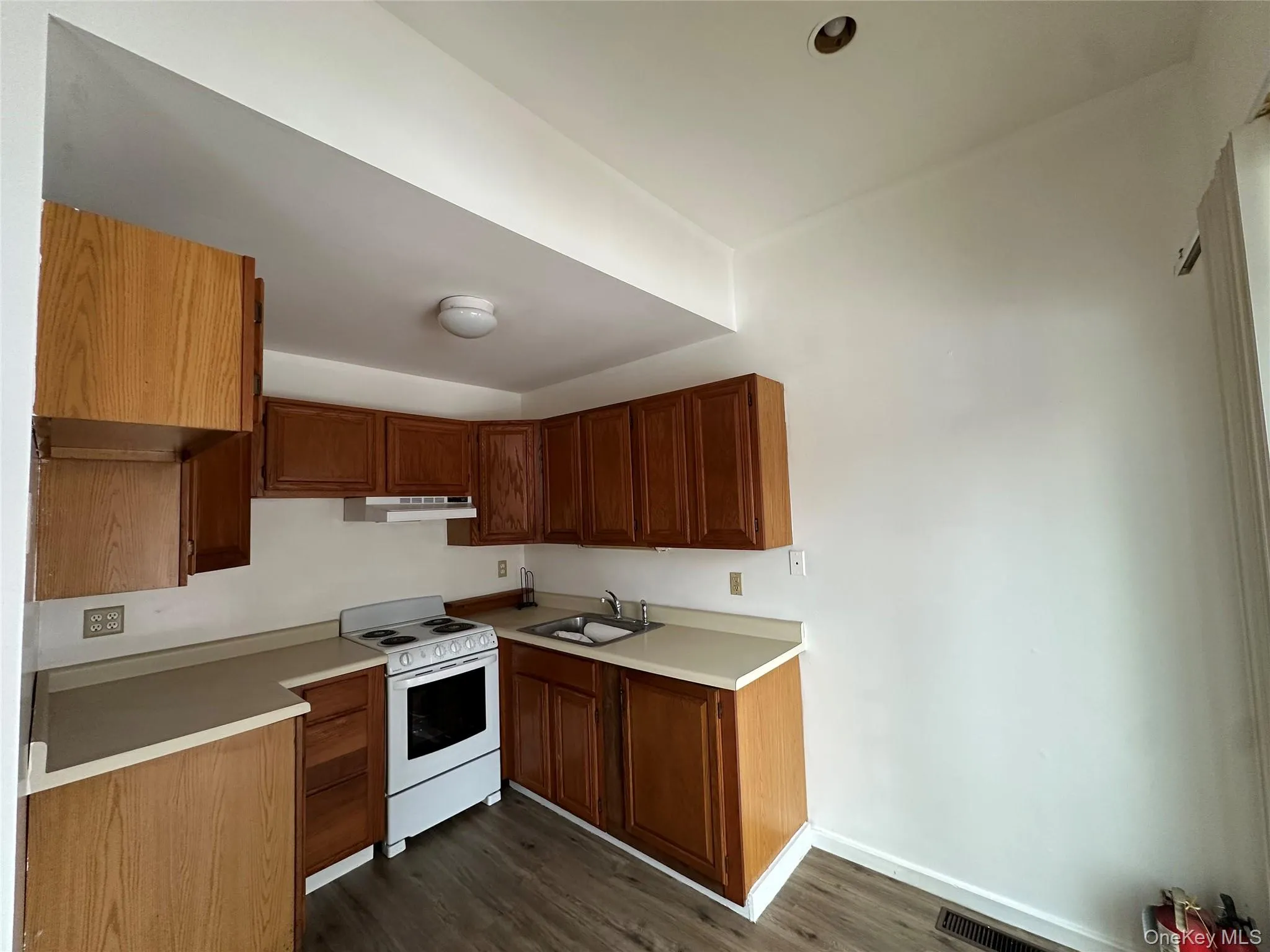 Kitchen with white range with electric stovetop, light countertops, under cabinet range hood, dark wood-style flooring, and brown cabinetry Kitchen with white range with electric stovetop, light countertops, under cabinet range hood, dark wood-style flooring, and brown cabinetry