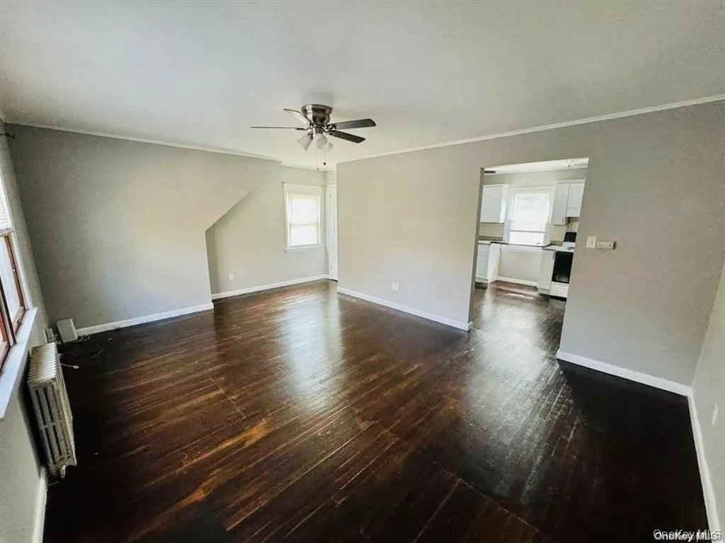 Empty room with radiator heating unit, plenty of natural light, crown molding, dark wood-style flooring, and a ceiling fan Empty room with radiator heating unit, plenty of natural light, crown molding, dark wood-style flooring, and a ceiling fan