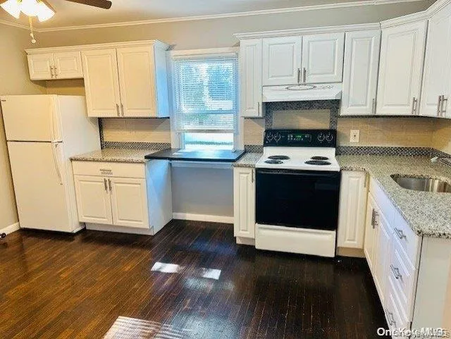 Kitchen featuring white appliances, crown molding, white cabinetry, dark wood-type flooring, and under cabinet range hood Kitchen featuring white appliances, crown molding, white cabinetry, dark wood-type flooring, and under cabinet range hood
