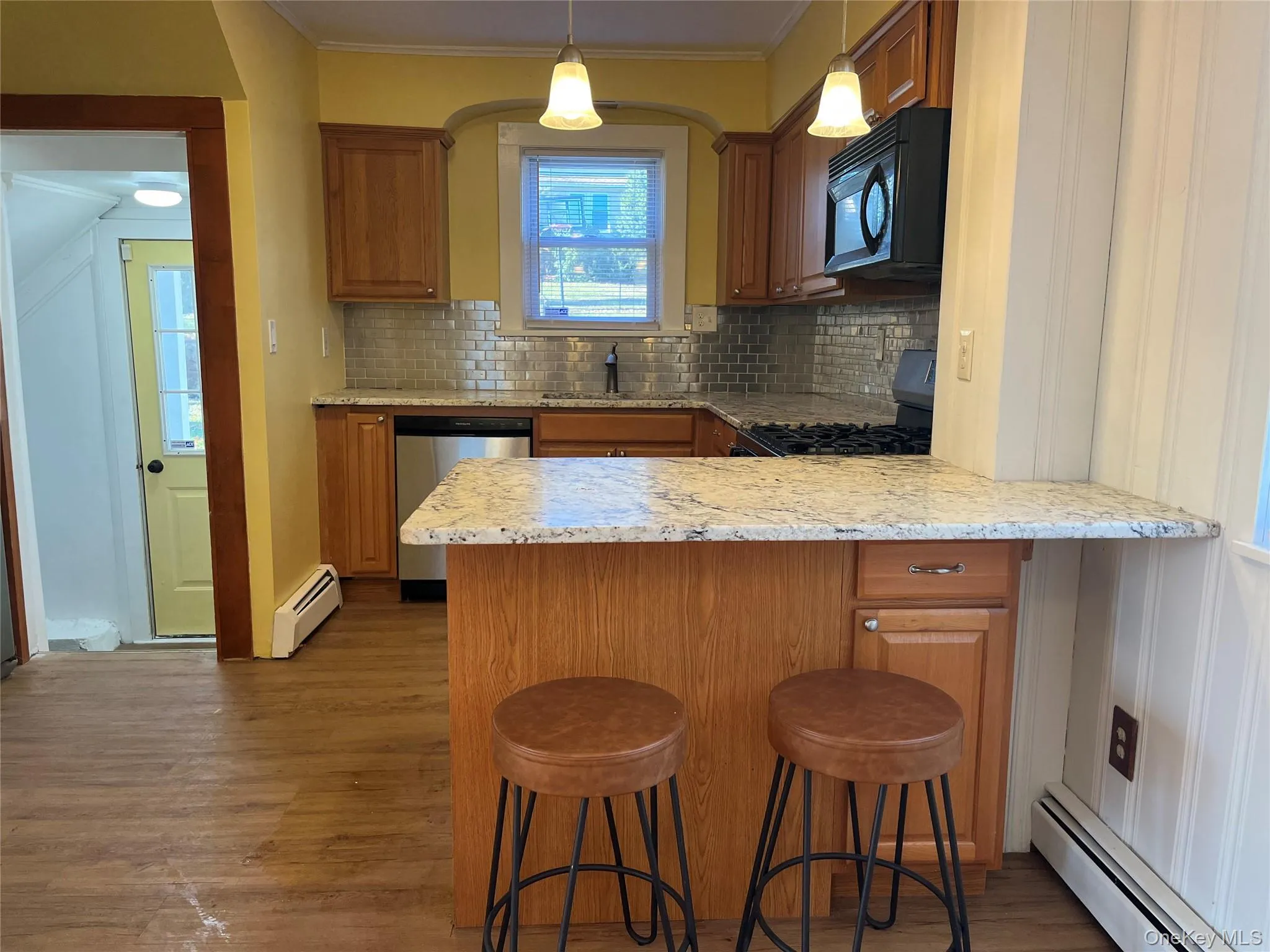 Kitchen with a baseboard radiator, a kitchen bar, a peninsula, black appliances, and brown cabinetry Kitchen with a baseboard radiator, a kitchen bar, a peninsula, black appliances, and brown cabinetry