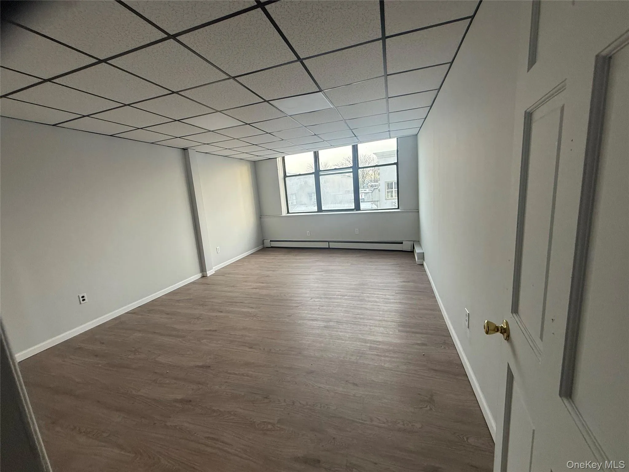 Empty room featuring a drop ceiling, dark wood-type flooring, and a baseboard heating unit Empty room featuring a drop ceiling, dark wood-type flooring, and a baseboard heating unit