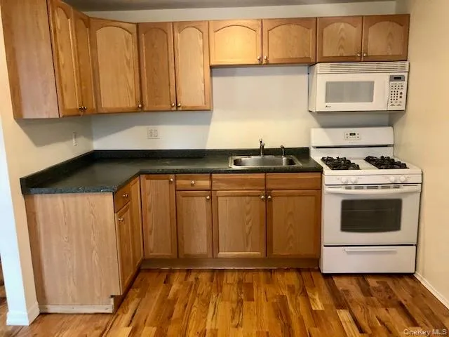 Kitchen with white appliances, dark countertops, dark wood finished floors, and brown cabinetry Kitchen with white appliances, dark countertops, dark wood finished floors, and brown cabinetry