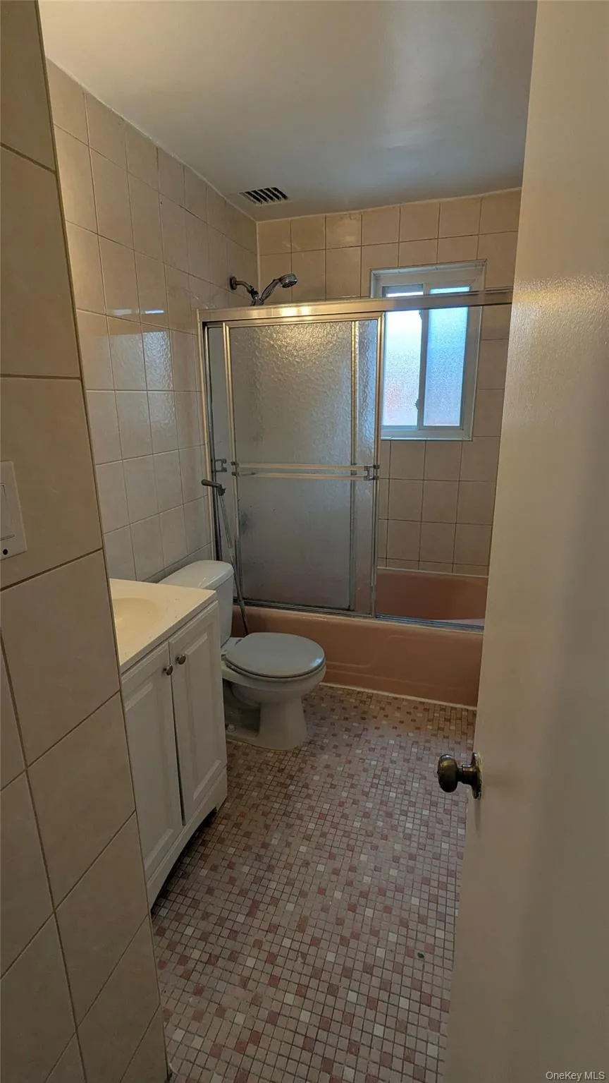 Bathroom featuring vanity, tile walls, shower / bath combination with glass door, and light tile patterned floors Bathroom featuring vanity, tile walls, shower / bath combination with glass door, and light tile patterned floors