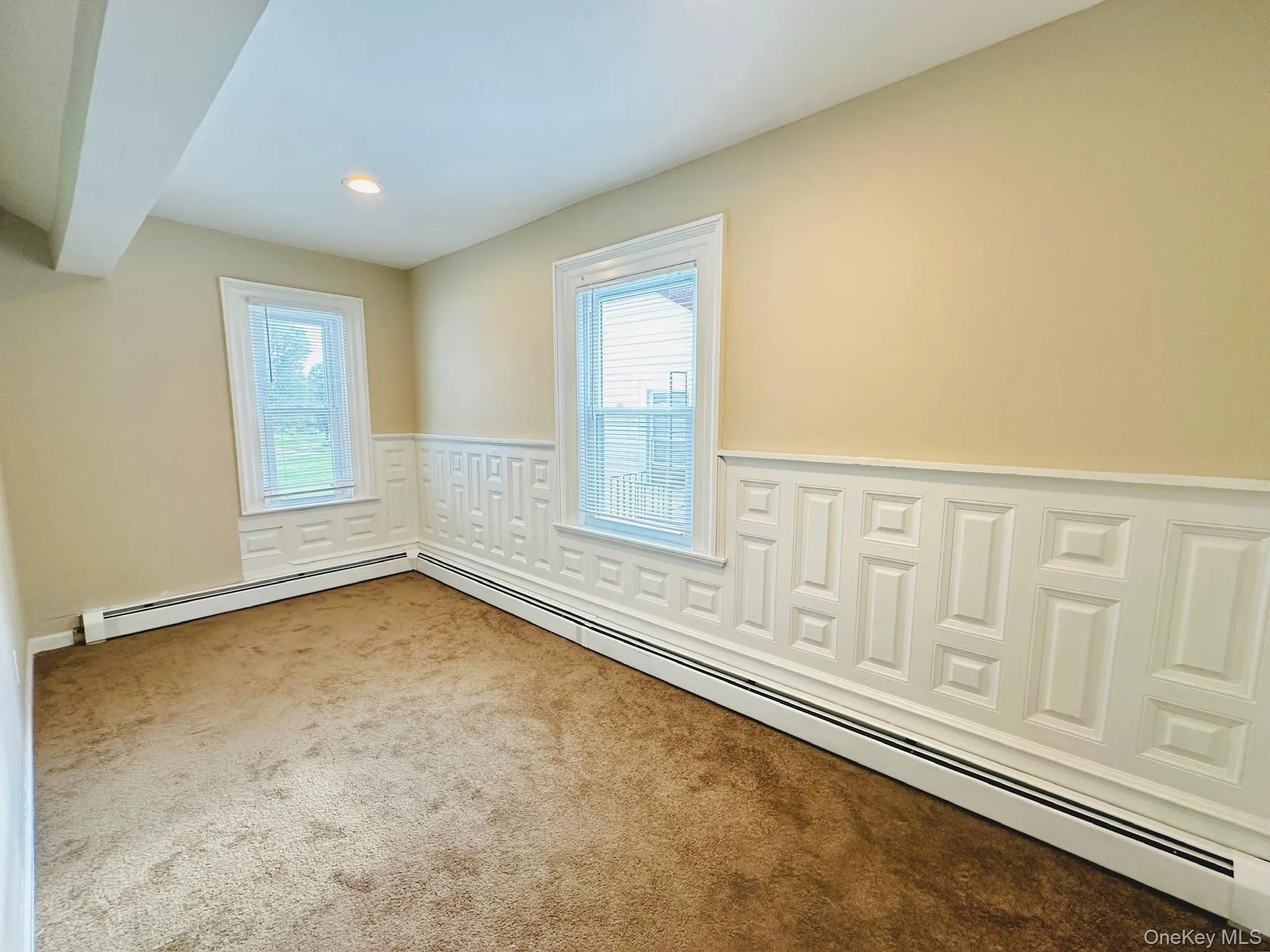 Empty room with baseboard heating, carpet, a wainscoted wall, recessed lighting, and a decorative wall Empty room with baseboard heating, carpet, a wainscoted wall, recessed lighting, and a decorative wall
