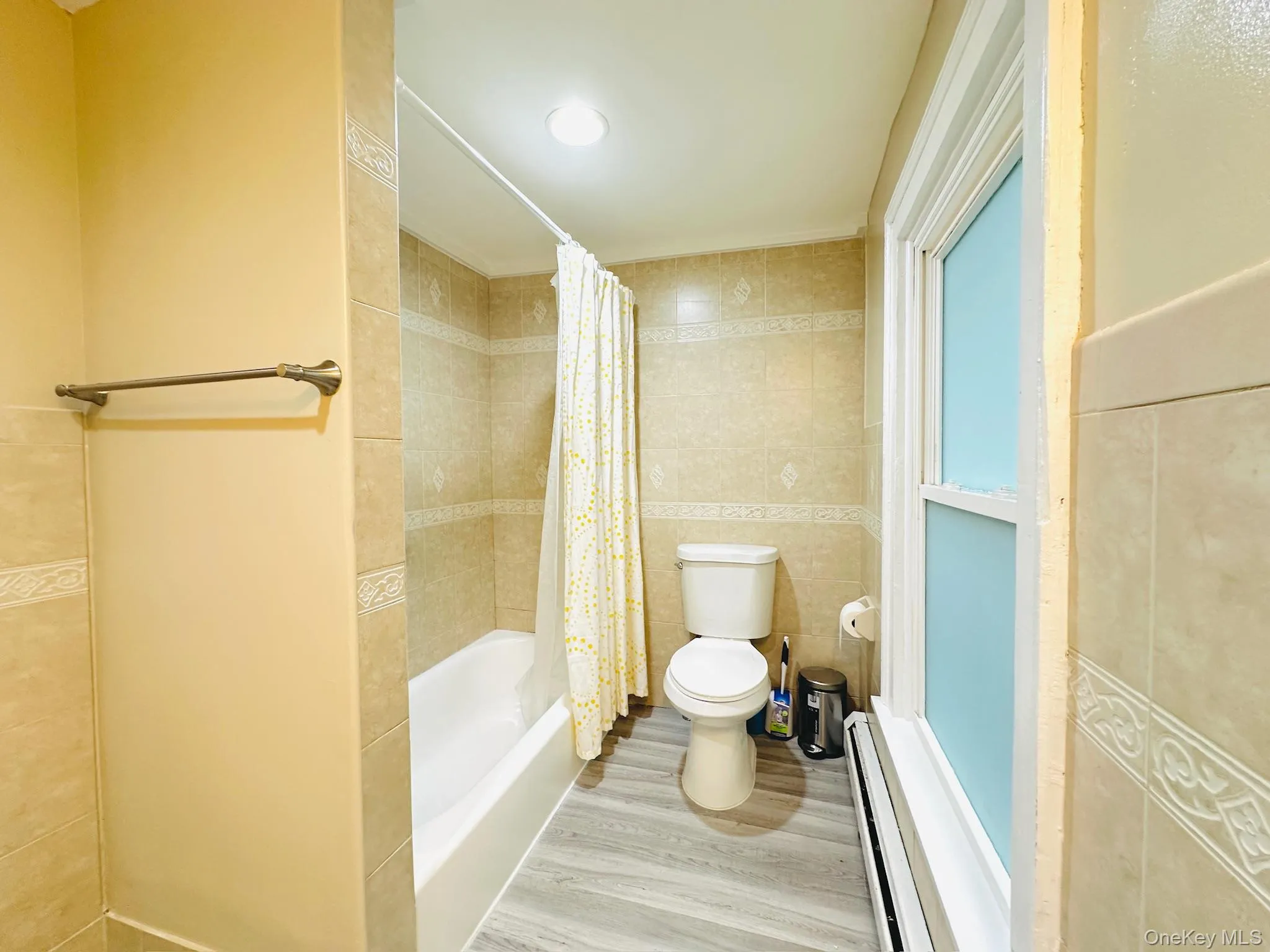 Bathroom with tile walls, baseboard heating, shower / tub combo with curtain, and wood finished floors Bathroom with tile walls, baseboard heating, shower / tub combo with curtain, and wood finished floors