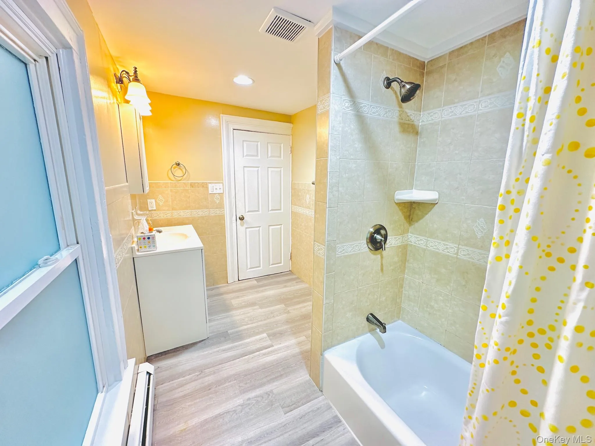 Full bathroom with tile walls, shower / bath combo, vanity, wainscoting, and a baseboard heating unit Full bathroom with tile walls, shower / bath combo, vanity, wainscoting, and a baseboard heating unit