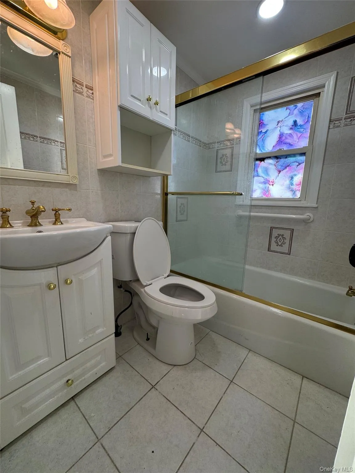 Full bath with vanity, bath / shower combo with glass door, tile walls, and light tile patterned floors Full bath with vanity, bath / shower combo with glass door, tile walls, and light tile patterned floors