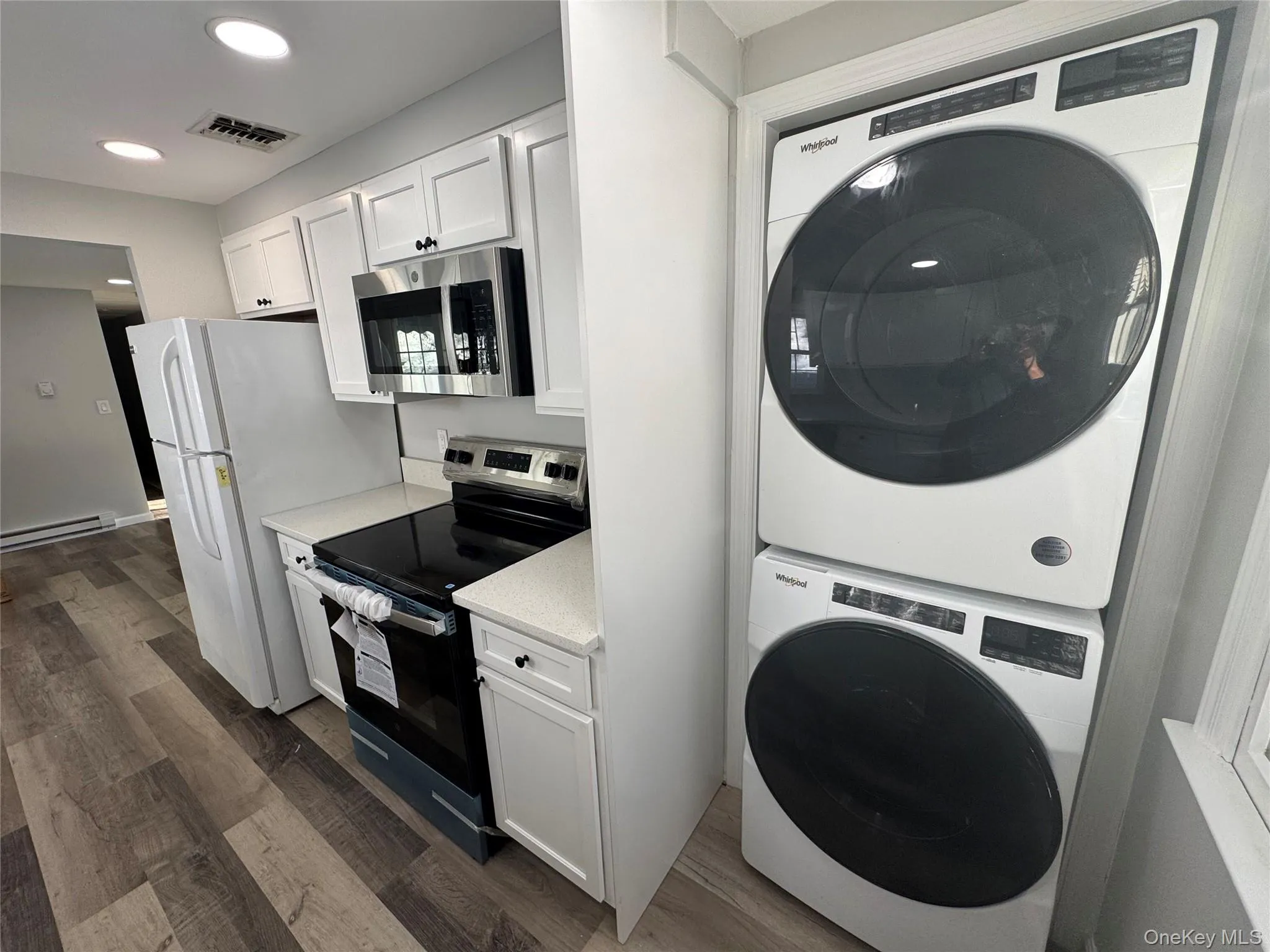 Kitchen featuring electric range oven, white cabinetry, stainless steel microwave, stacked washer / dryer, and dark wood-style floors Kitchen featuring electric range oven, white cabinetry, stainless steel microwave, stacked washer / dryer, and dark wood-style floors