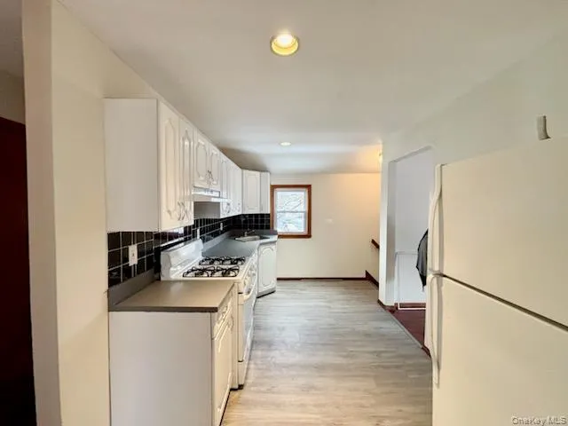Kitchen with white appliances, dark countertops, light wood-style flooring, backsplash, and white cabinetry Kitchen with white appliances, dark countertops, light wood-style flooring, backsplash, and white cabinetry
