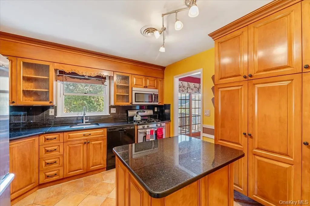 Kitchen featuring tasteful backsplash, appliances with stainless steel finishes, a center island, brown cabinetry, and dark stone countertops Kitchen featuring tasteful backsplash, appliances with stainless steel finishes, a center island, brown cabinetry, and dark stone countertops