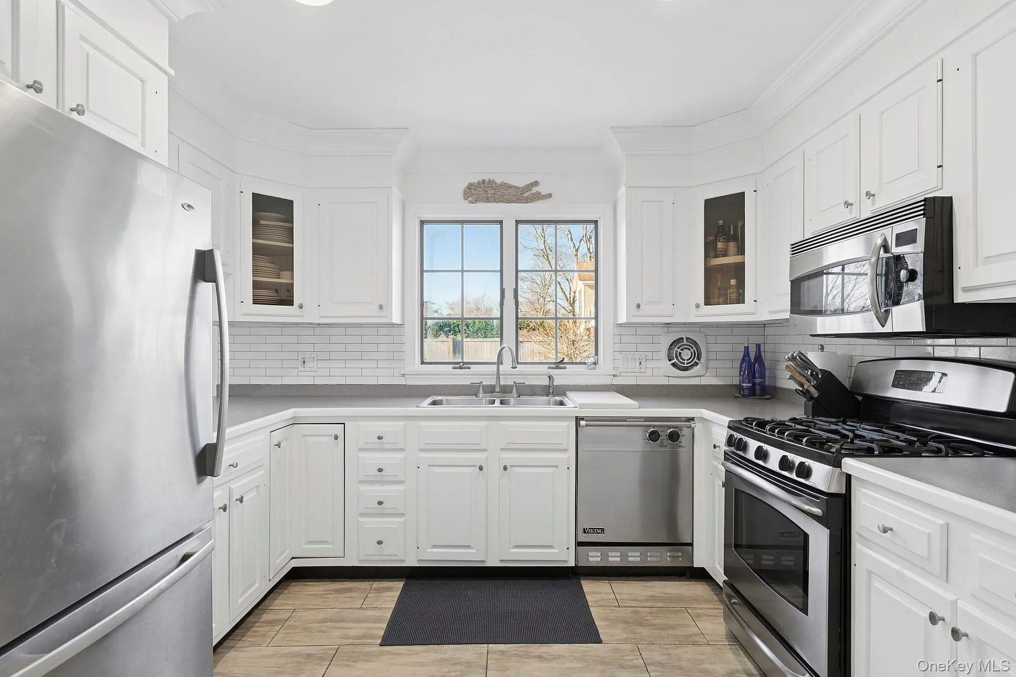 Kitchen featuring appliances with stainless steel finishes, white cabinetry, glass insert cabinets, ornamental molding, and light countertops Kitchen featuring appliances with stainless steel finishes, white cabinetry, glass insert cabinets, ornamental molding, and light countertops