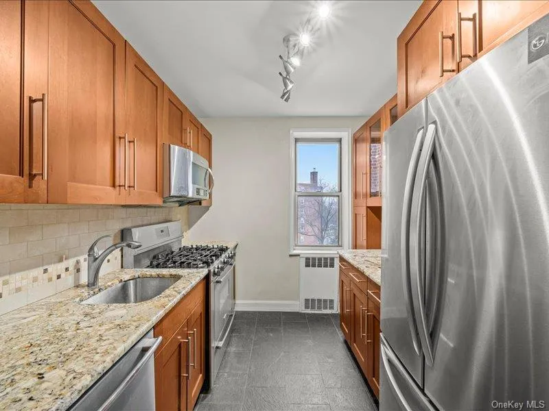 Kitchen with stainless steel appliances, light stone counters, brown cabinetry, and radiator heating unit Kitchen with stainless steel appliances, light stone counters, brown cabinetry, and radiator heating unit