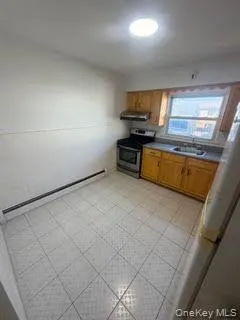 Kitchen with stainless steel range oven, a baseboard radiator, brown cabinetry, light flooring, and freestanding refrigerator Kitchen with stainless steel range oven, a baseboard radiator, brown cabinetry, light flooring, and freestanding refrigerator