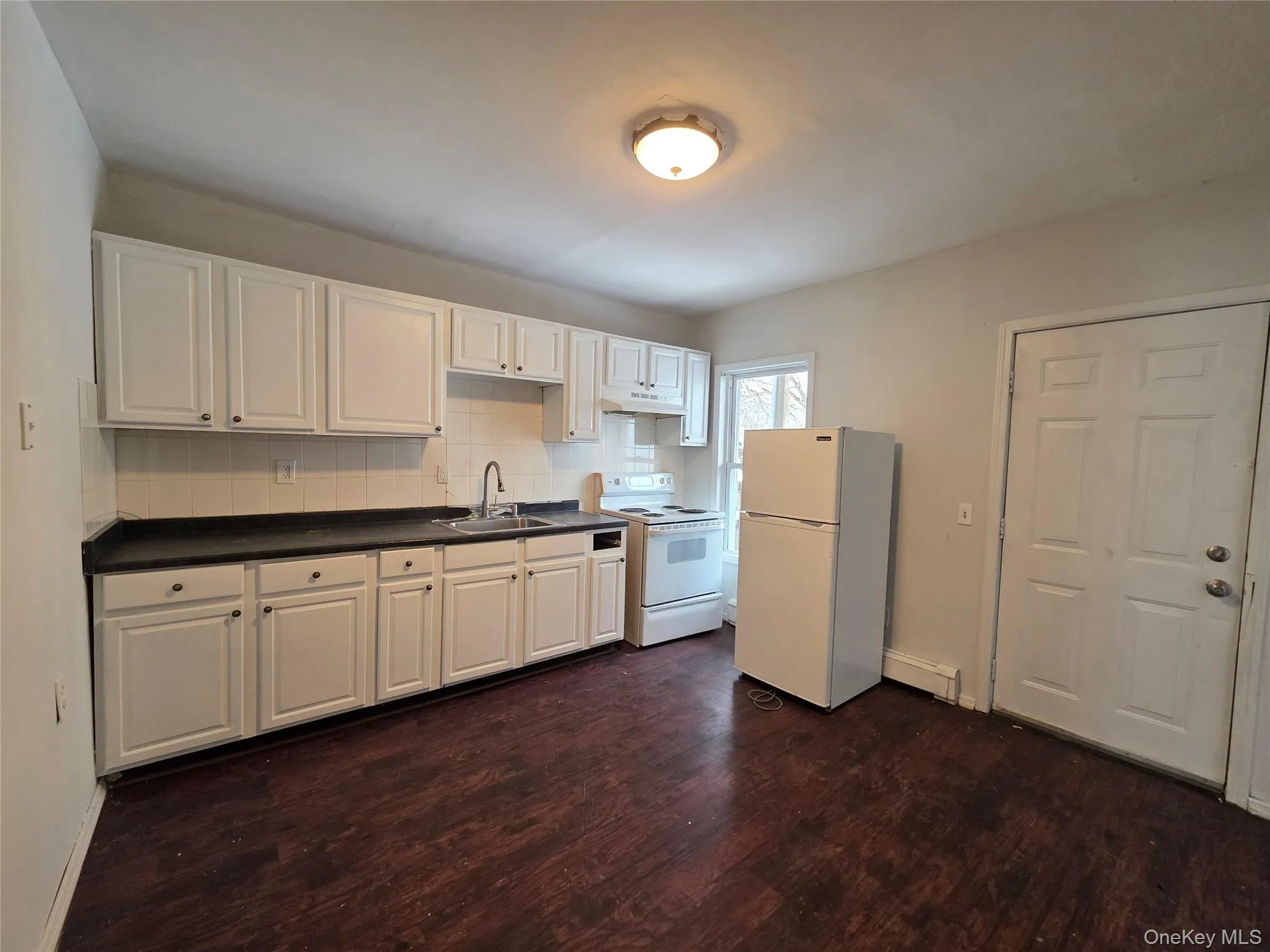 Kitchen with white appliances, white cabinetry, dark countertops, tasteful backsplash, and dark wood finished floors Kitchen with white appliances, white cabinetry, dark countertops, tasteful backsplash, and dark wood finished floors